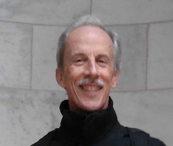 An elderly man with gray hair and a mustache smiling, wearing a black jacket, standing against a gray stone wall.