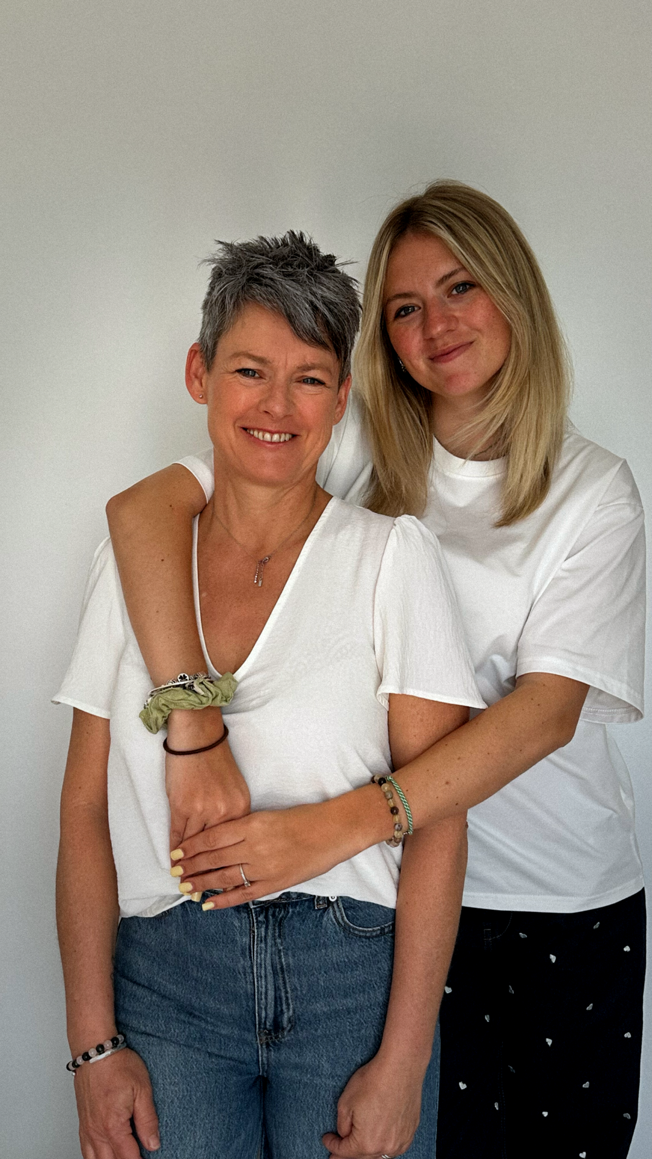 Deux femmes souriantes, l'une avec des cheveux courts et gris et l'autre avec des cheveux longs blonds, posent contre un mur blanc, la femme plus âgée a l'air d'être la mère ou une mère et la plus jeune une fille ou une amie.
