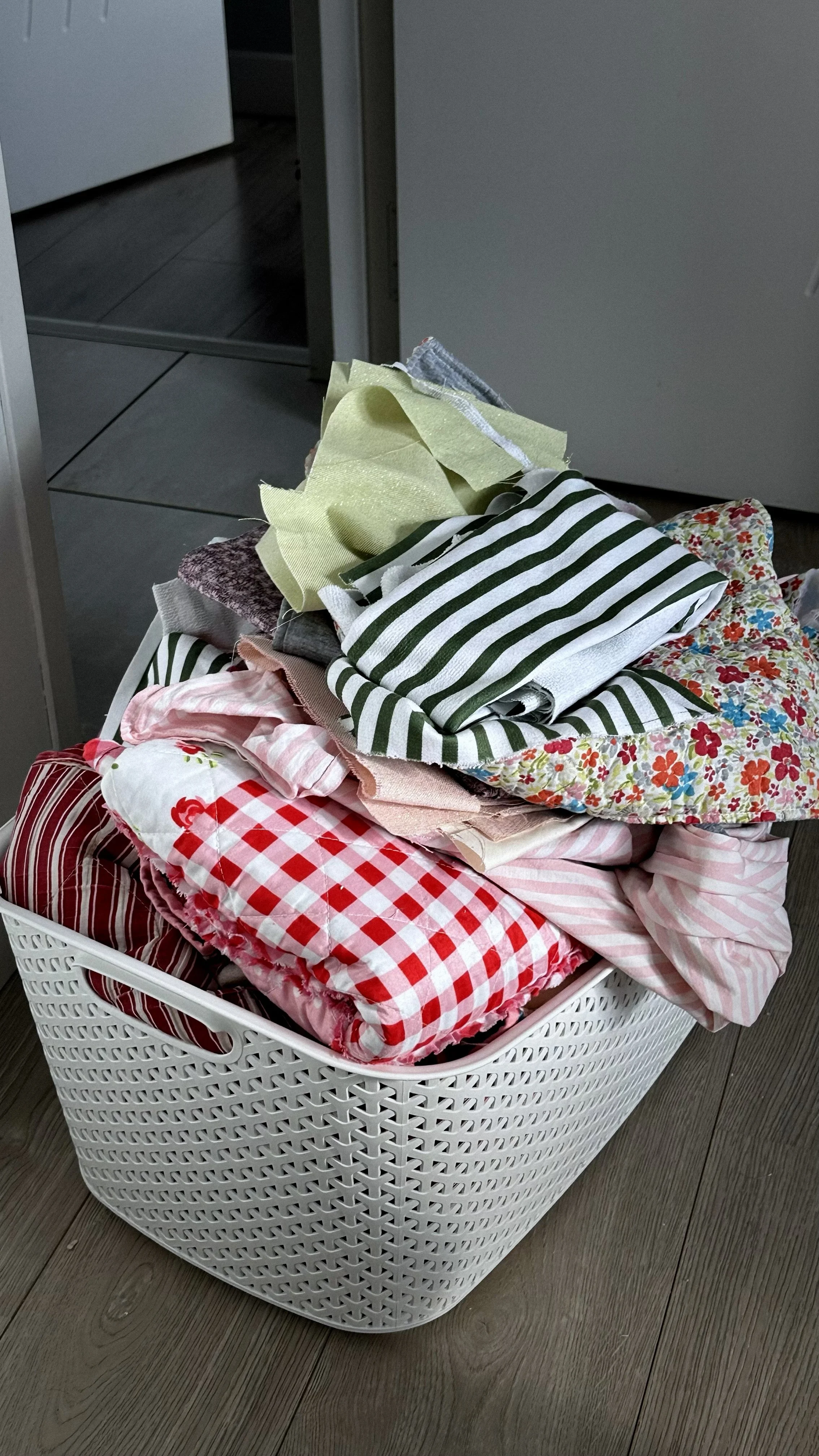 Panier en plastique blanc rempli de linges ou serviettes colorés et à motifs, placé sur un sol en bois.