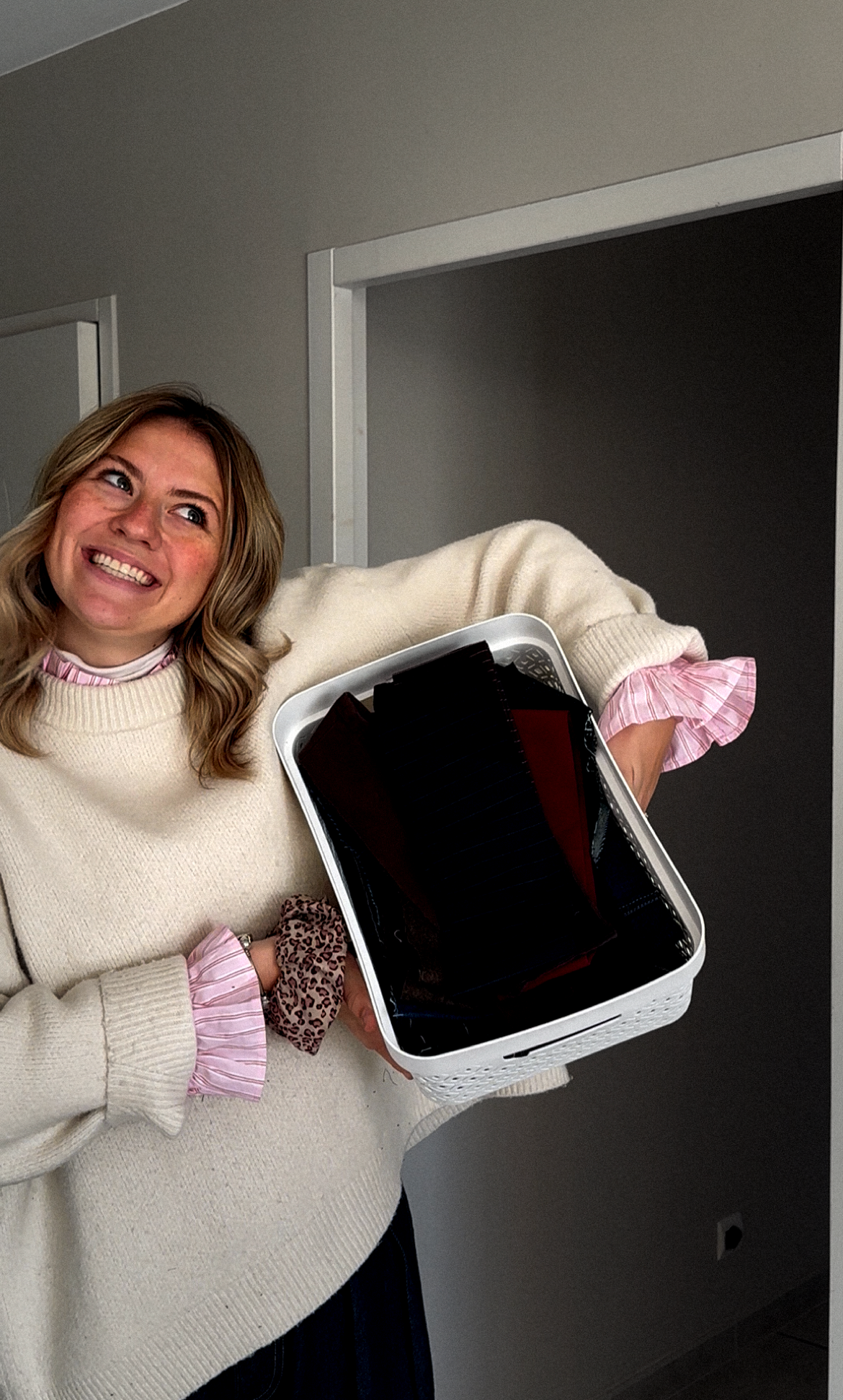 Une femme souriante tient une boîte blanche contenant des dossiers ou des papiers en plastique noirs. Elle porte un pull beige avec des manches visibles rose à rayures.