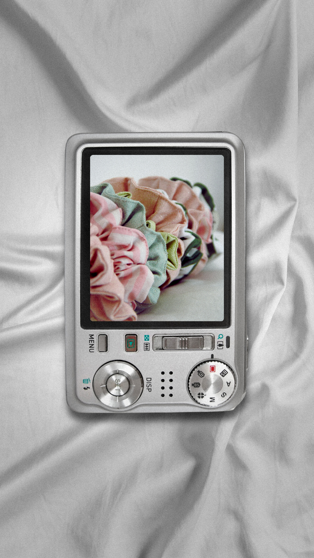 Téléphone portable silver affichant une photo de chiffons colorés sur son écran, posé sur un tissu blanc.