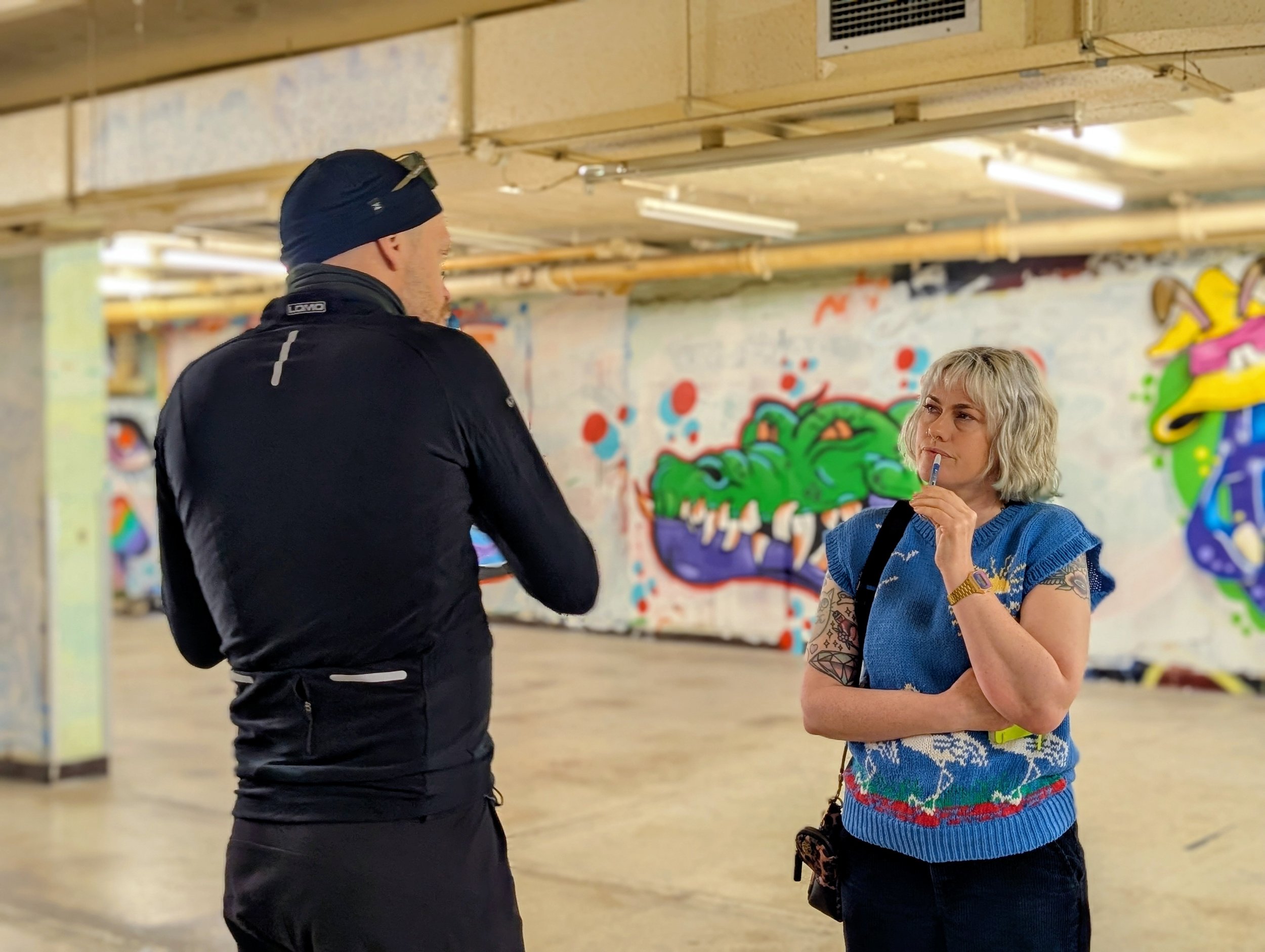 A man wearing a black jacket and a blue beanie hat is talking to a woman with curly gray hair, tattoos on her arms, and a blue sweater in an indoor space with colorful graffiti art on the walls.