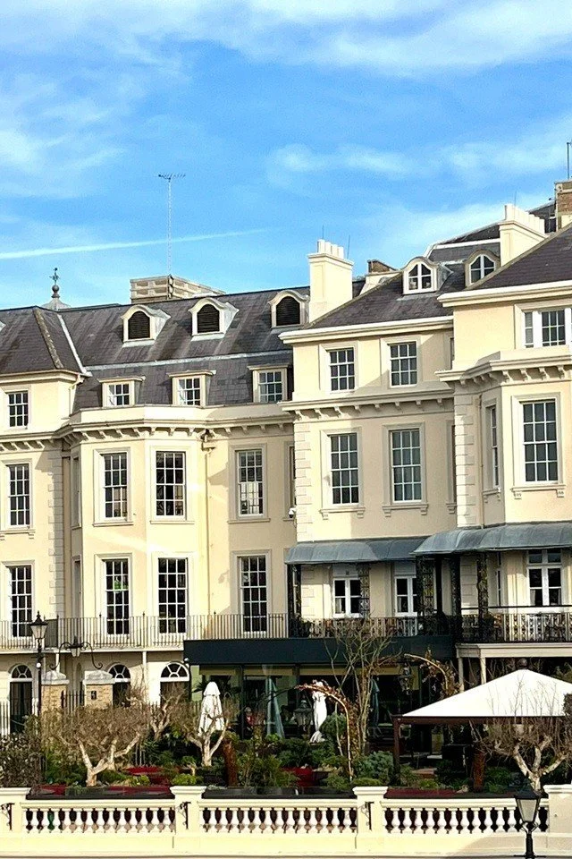 Elegant multi-story European-style building with large windows, balconies, and a courtyard with trees and outdoor seating under a blue sky.