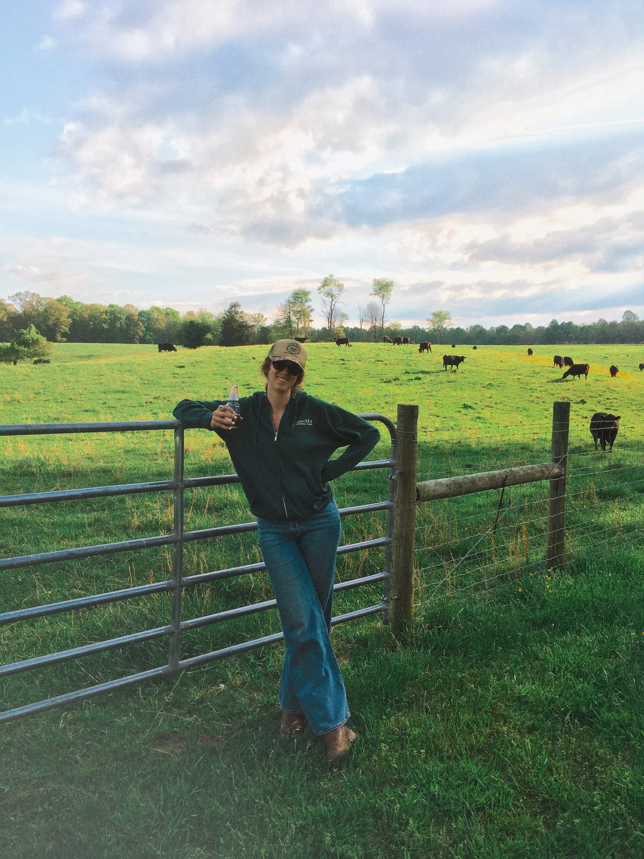 A woman standing by a metal fence with a green pasture, cows grazing, and trees with a partly cloudy sky in the background.