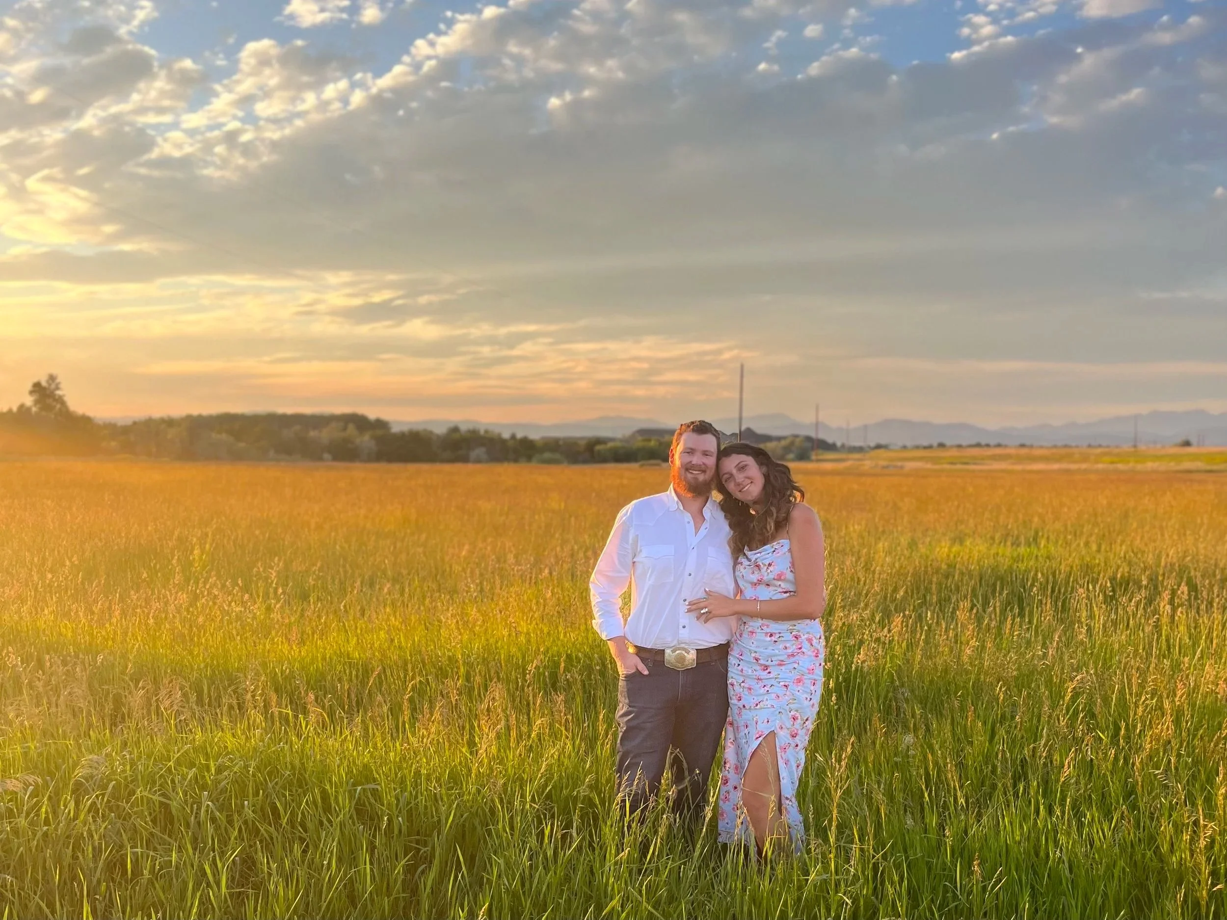 A happy couple standing in a golden field at sunset with a partly cloudy sky overhead.