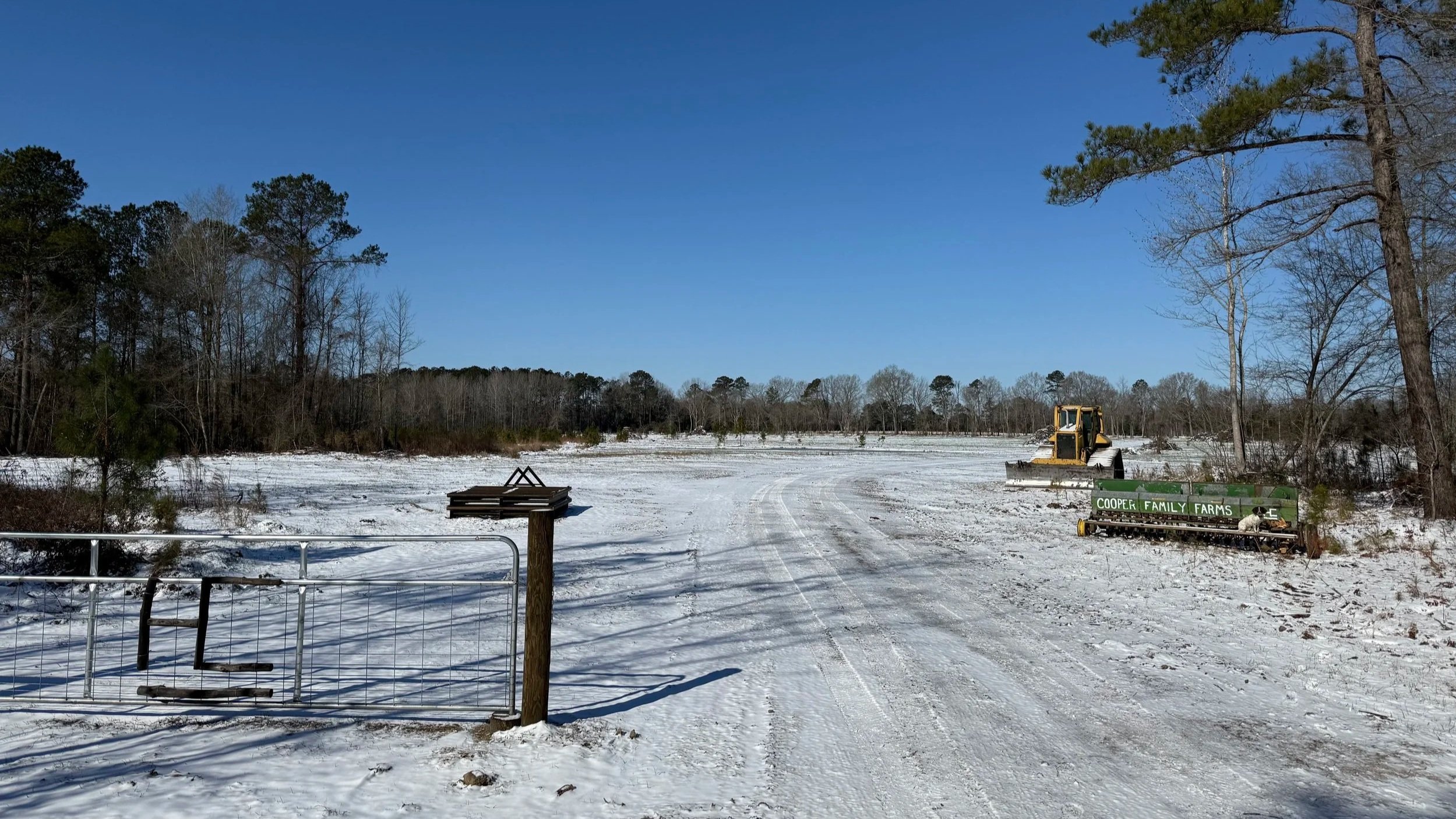 Snow-covered farm field with a yellow tractor and a green farm equipment trailer labeled "Cooper Family Farms" on the right, trees in the background, and a clear blue sky.