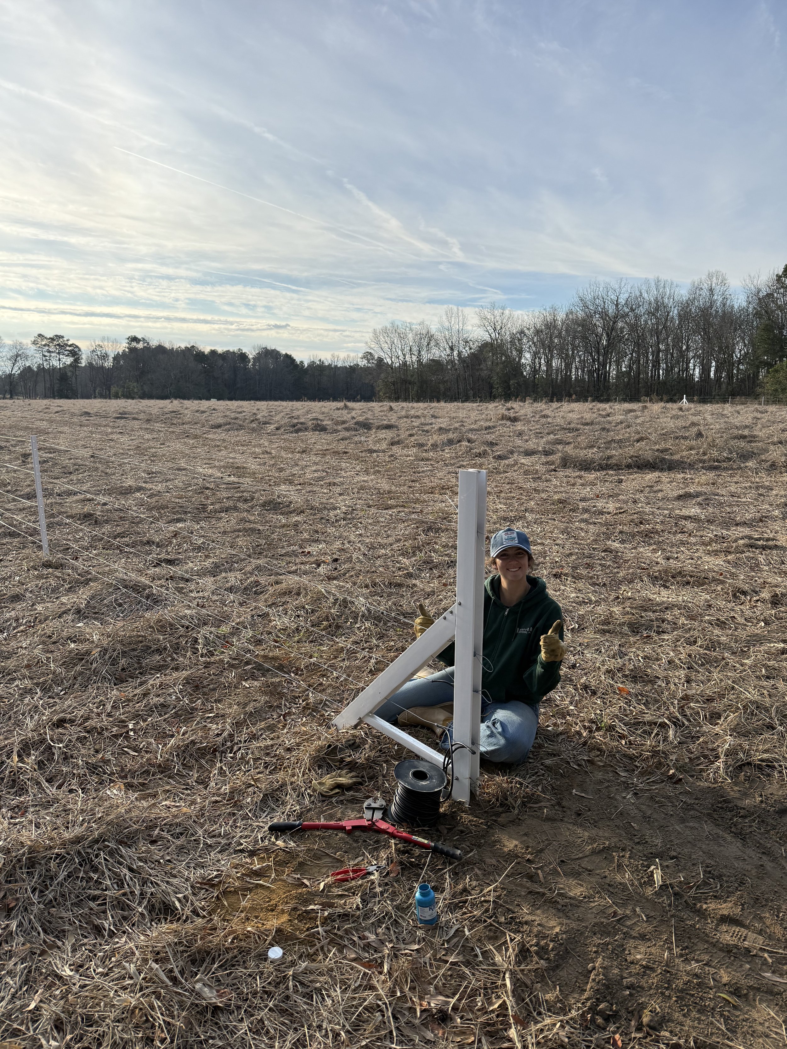 A woman is smiling and crouching in a field, working on a small satellite dish and equipment, with a partly cloudy sky and a line of trees in the background.