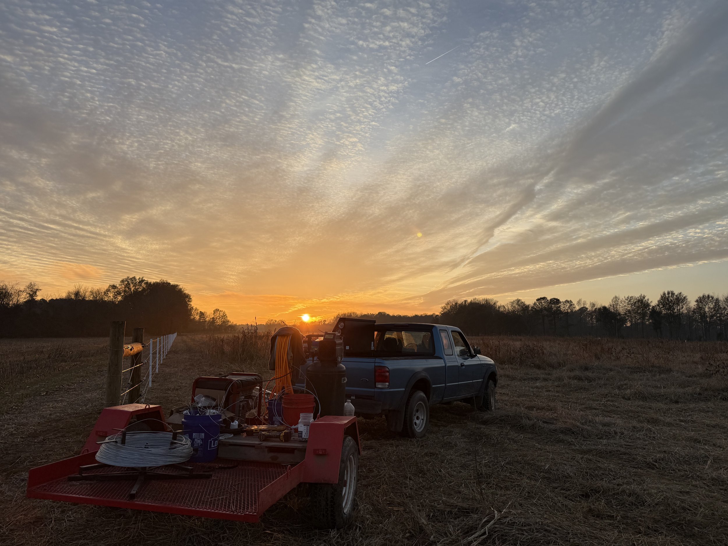 Sunset over a rural field with parked pickup truck, gardening tools, and supplies in foreground, with trees and an open sky with scattered clouds and a jet trail.