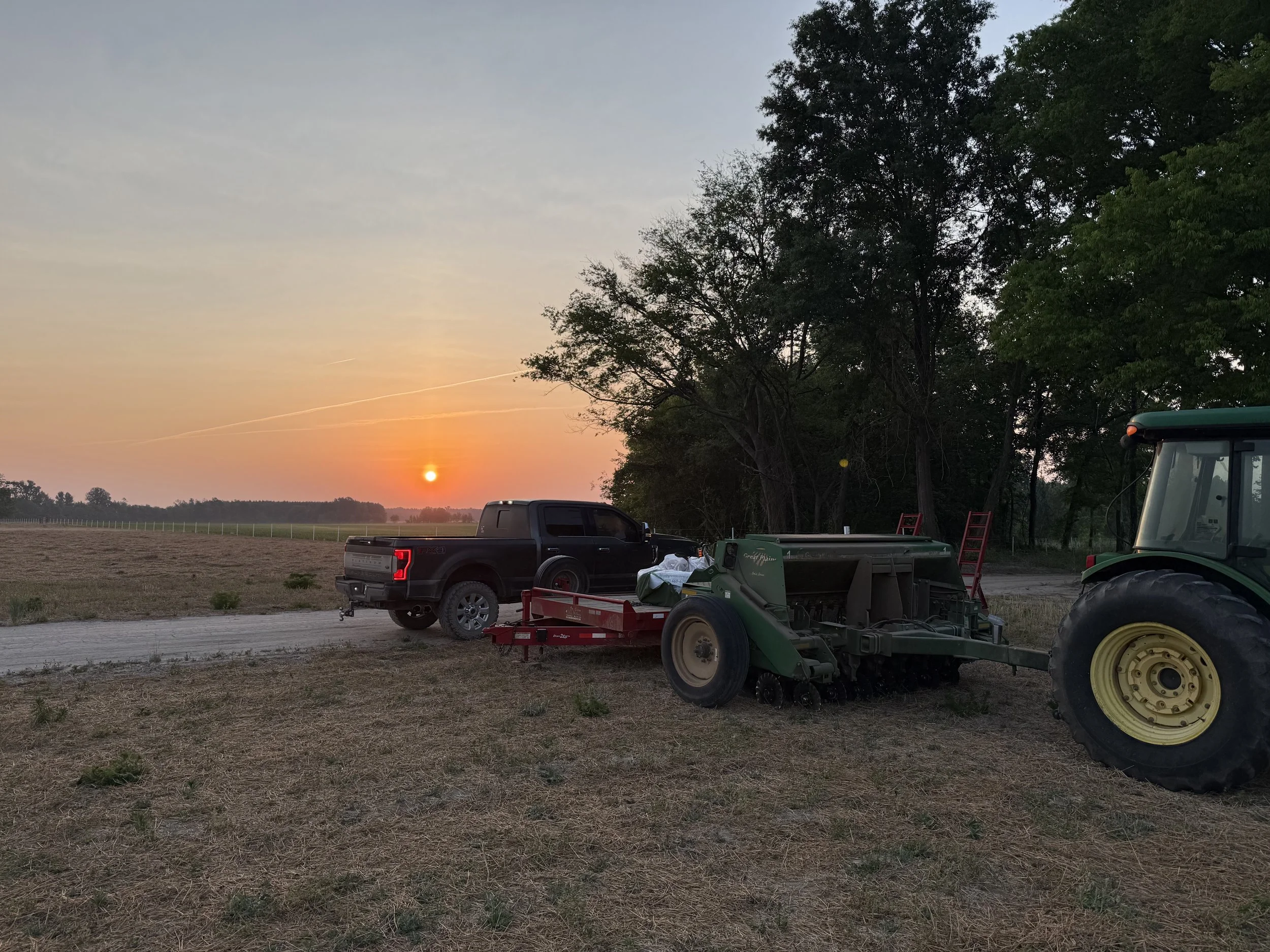 Sunset over a farm field with equipment, trucks, and trees in the background.