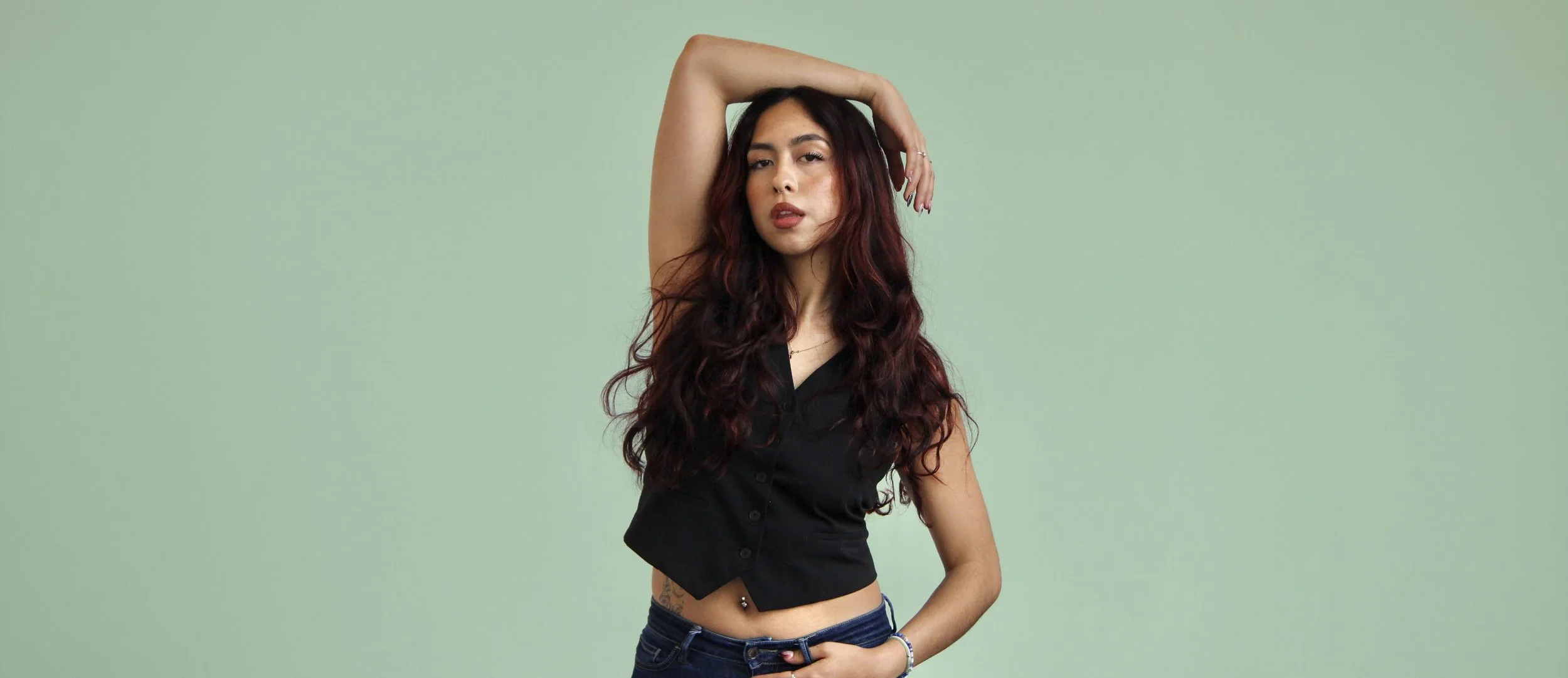 A young woman with long, curly red hair and a nose piercing poses with one arm raised above her head, wearing a black sleeveless shirt and blue jeans against a plain light green background.