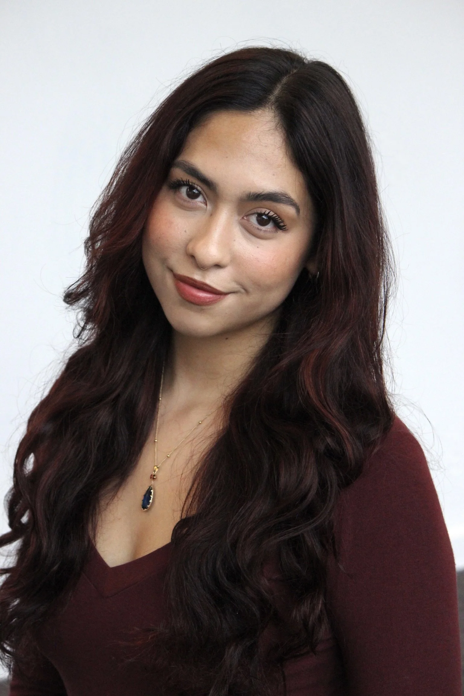A woman with long, wavy, dark red hair styled in loose curls, wearing a maroon top and a gold necklace with a blue pendant, smiling softly against a plain white background.