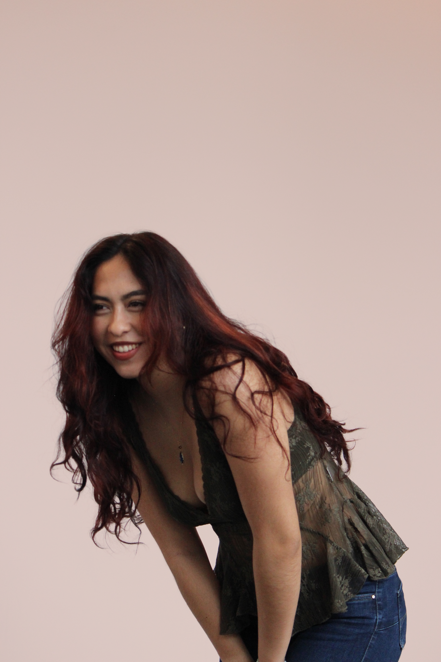 A woman with long, wavy, reddish-brown hair smiling and leaning forward against a plain, light-colored wall.