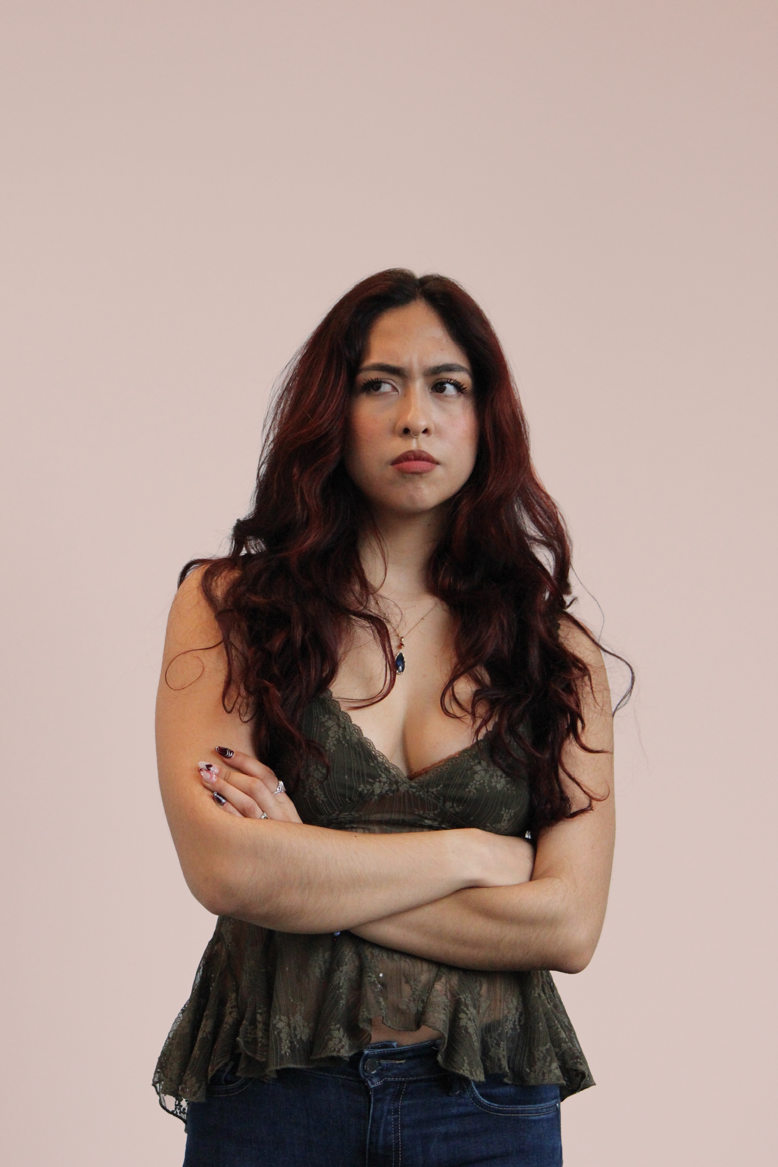 A woman with long, dark red, wavy hair standing against a light-colored background, crossing her arms and looking to her left with a serious expression.