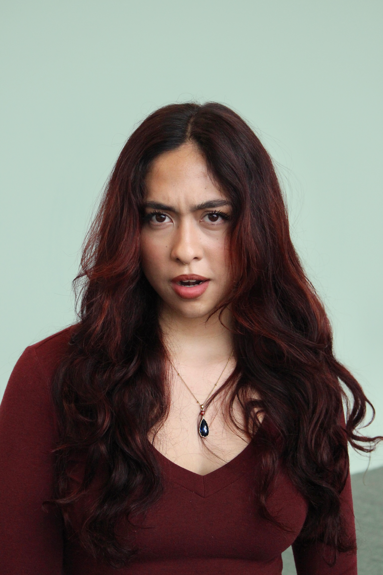 A woman with long, dark red hair and tan skin wearing a maroon top and a pendant necklace, looking at the camera with a surprised expression, against a pale mint green background.