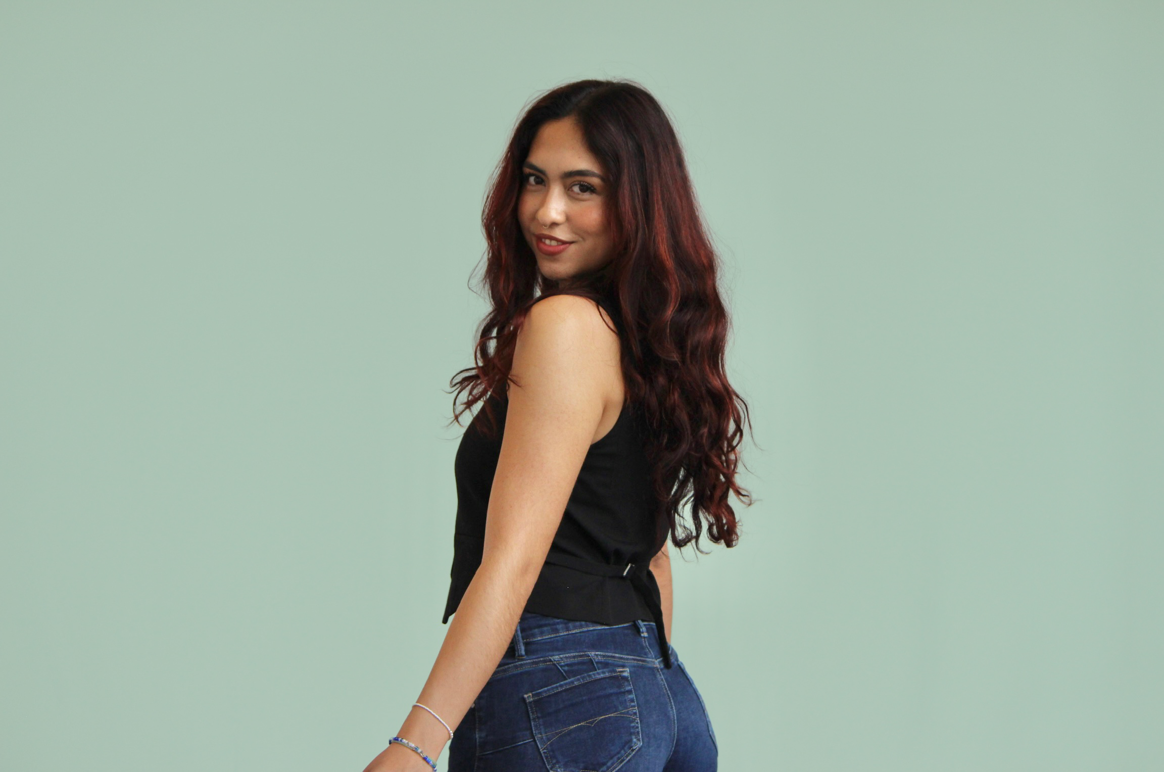 A young woman with long, wavy, dark red hair, posing in front of a light green background, wearing a sleeveless black top and blue jeans, smiling slightly while looking over her shoulder.