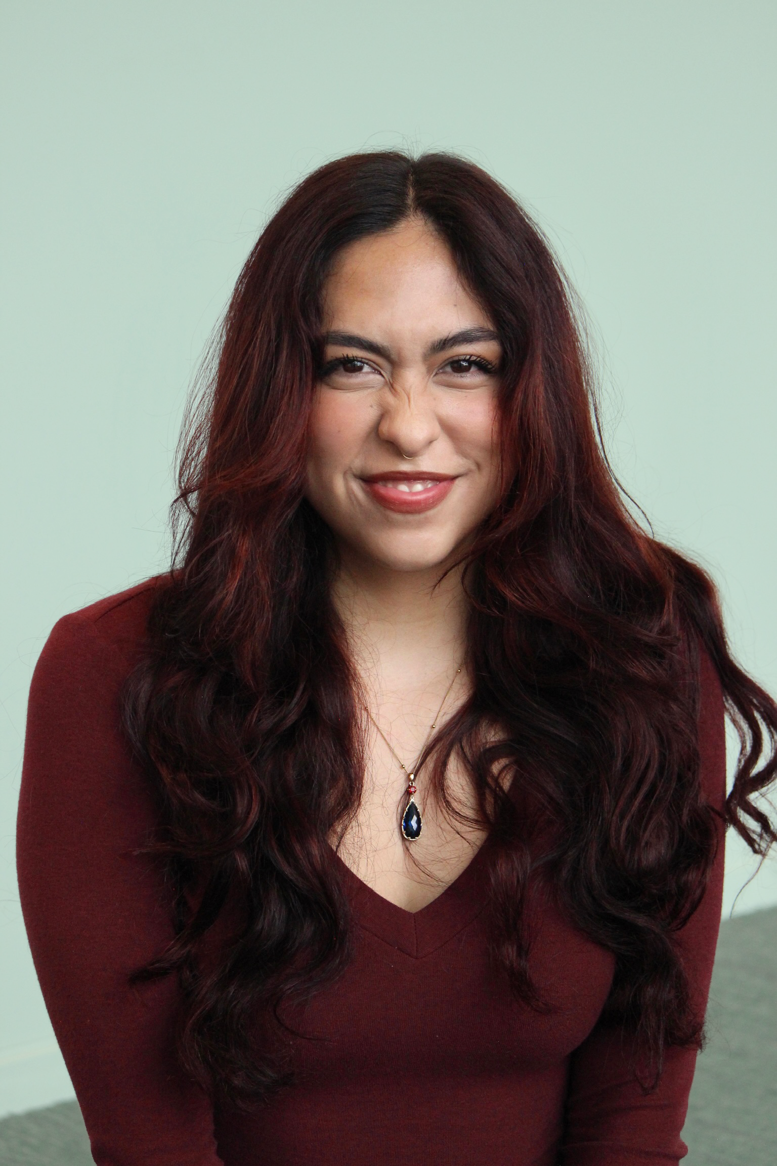 A woman with long, dark red, wavy hair smiling, wearing a maroon top and a blue pendant necklace, standing against a light green background.