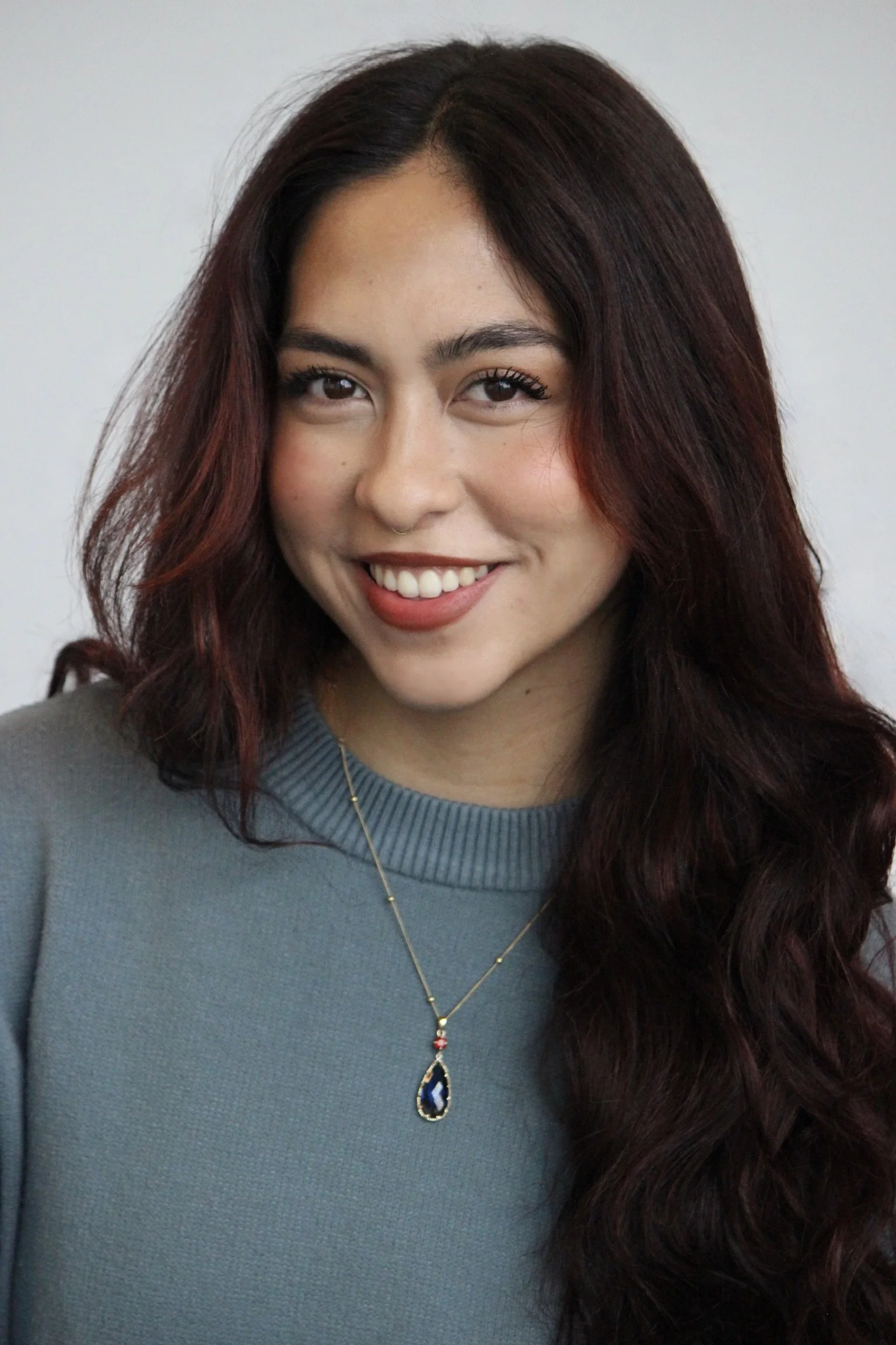 A woman with long, dark auburn hair, smiling, wearing a gray top and a jewelry necklace with a blue gemstone pendant.