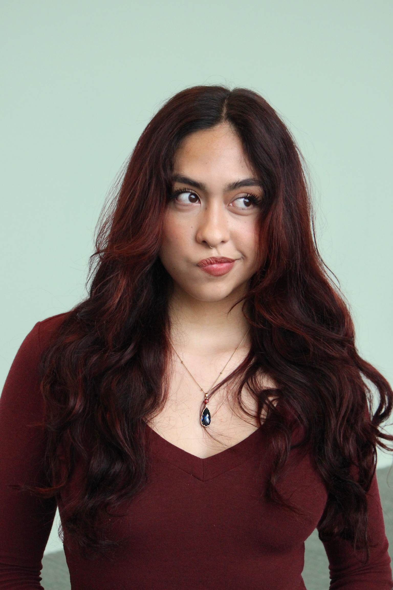 A woman with long, wavy, burgundy hair wearing a burgundy top and a pendant necklace, looking to her right with a neutral expression, standing against a light green background.