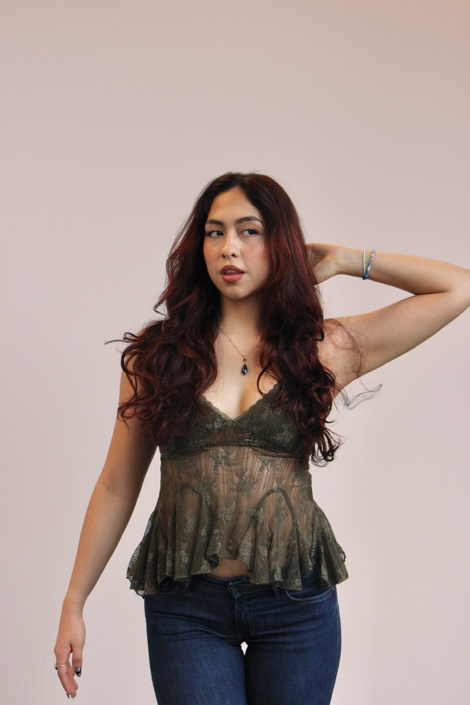 A woman with long, wavy dark red hair posing against a plain wall, wearing a sheer, patterned brown top and dark blue jeans, with one hand behind her head and the other relaxed by her side, accessorized with jewelry.