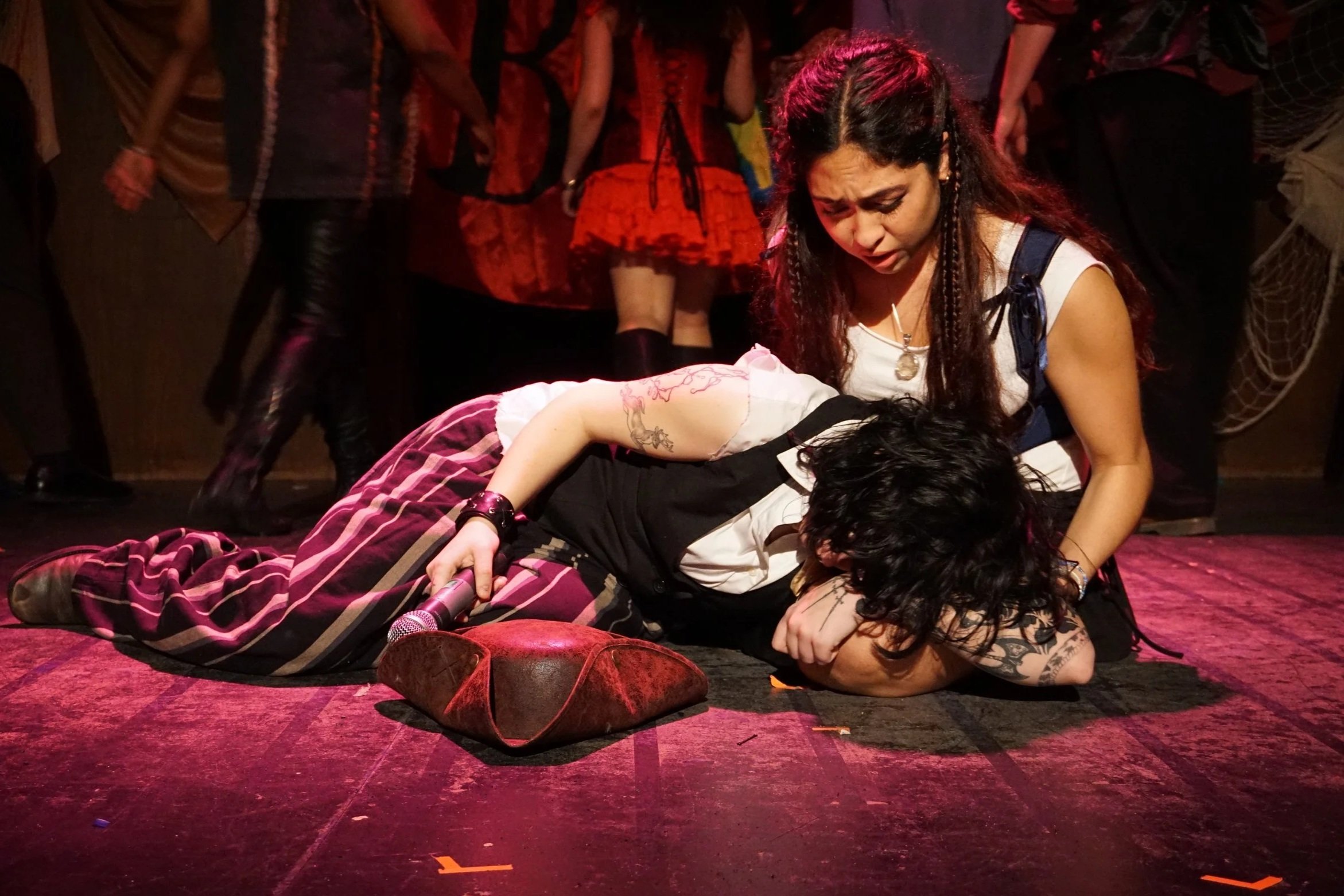 A woman with long dark hair and tattoos on her arm, wearing a white top, kneels over a shirtless person lying on the ground, holding a microphone and appearing upset. Several people stand around in the background, wearing dark and colorful clothing, on a stage with red and purple lighting.