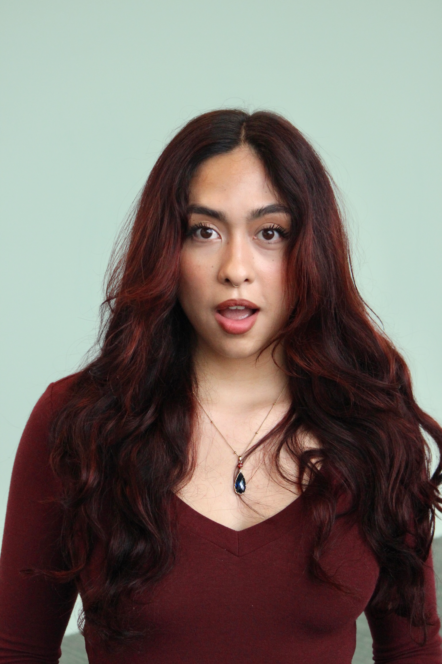 A woman with long, wavy, dark red hair, wearing a maroon top and a necklace with a teardrop-shaped pendant, standing against a light-colored background.