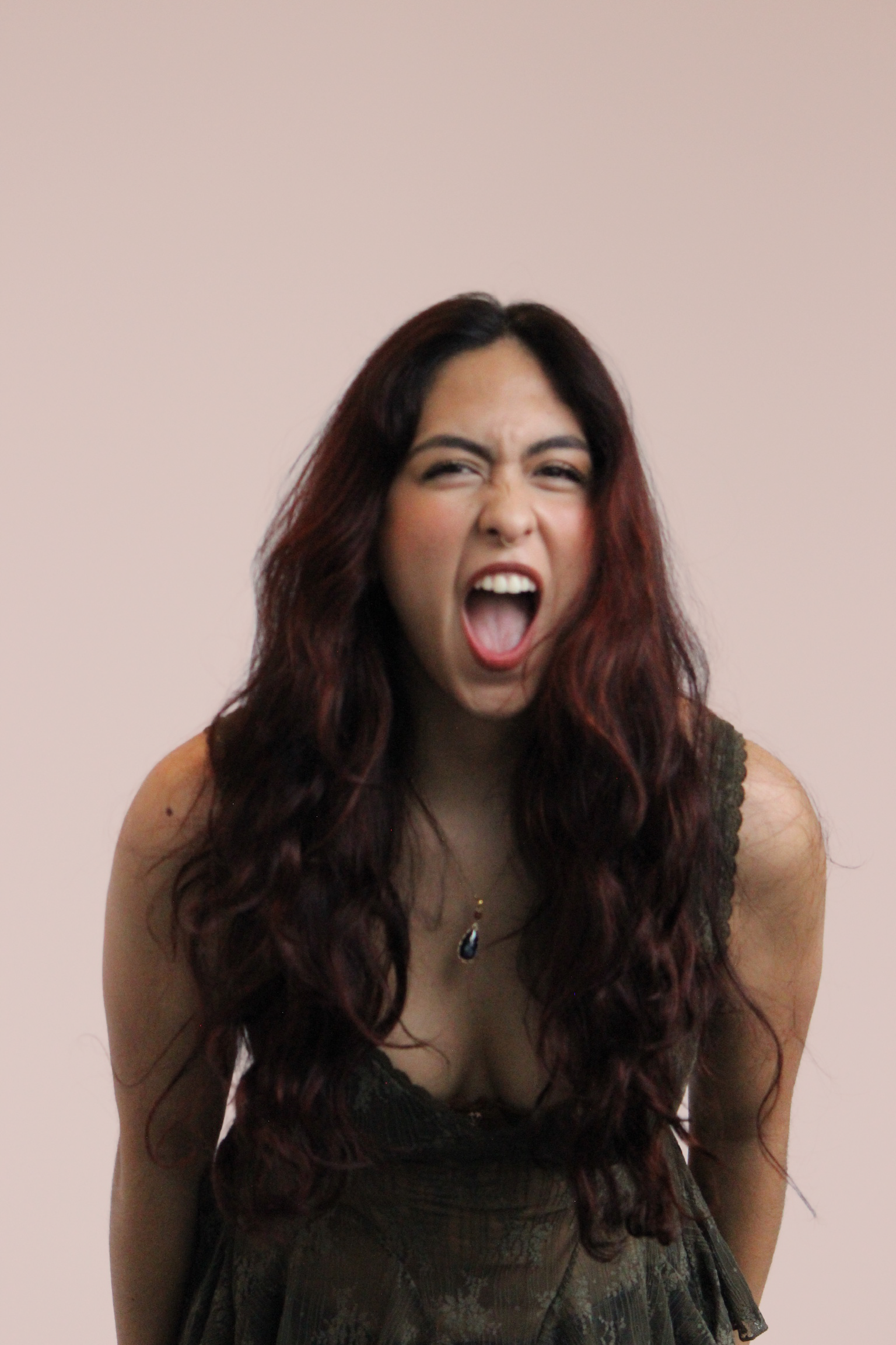 Young woman with long wavy dark red hair, mid-yawn or shout, wearing a sleeveless dark dress and a necklace with a dark pendant, standing against a plain light-colored background.