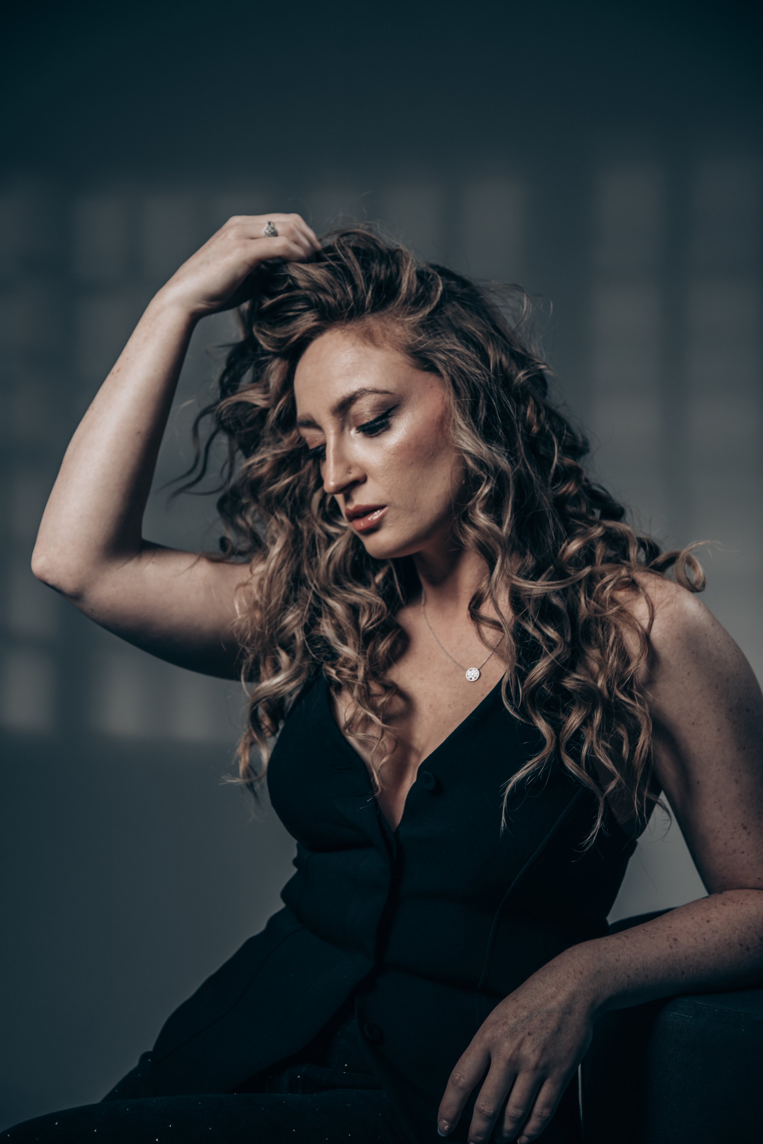 A woman with curly hair and light skin wearing a black top, looking down with her hand on her head, in a dimly lit indoor setting.