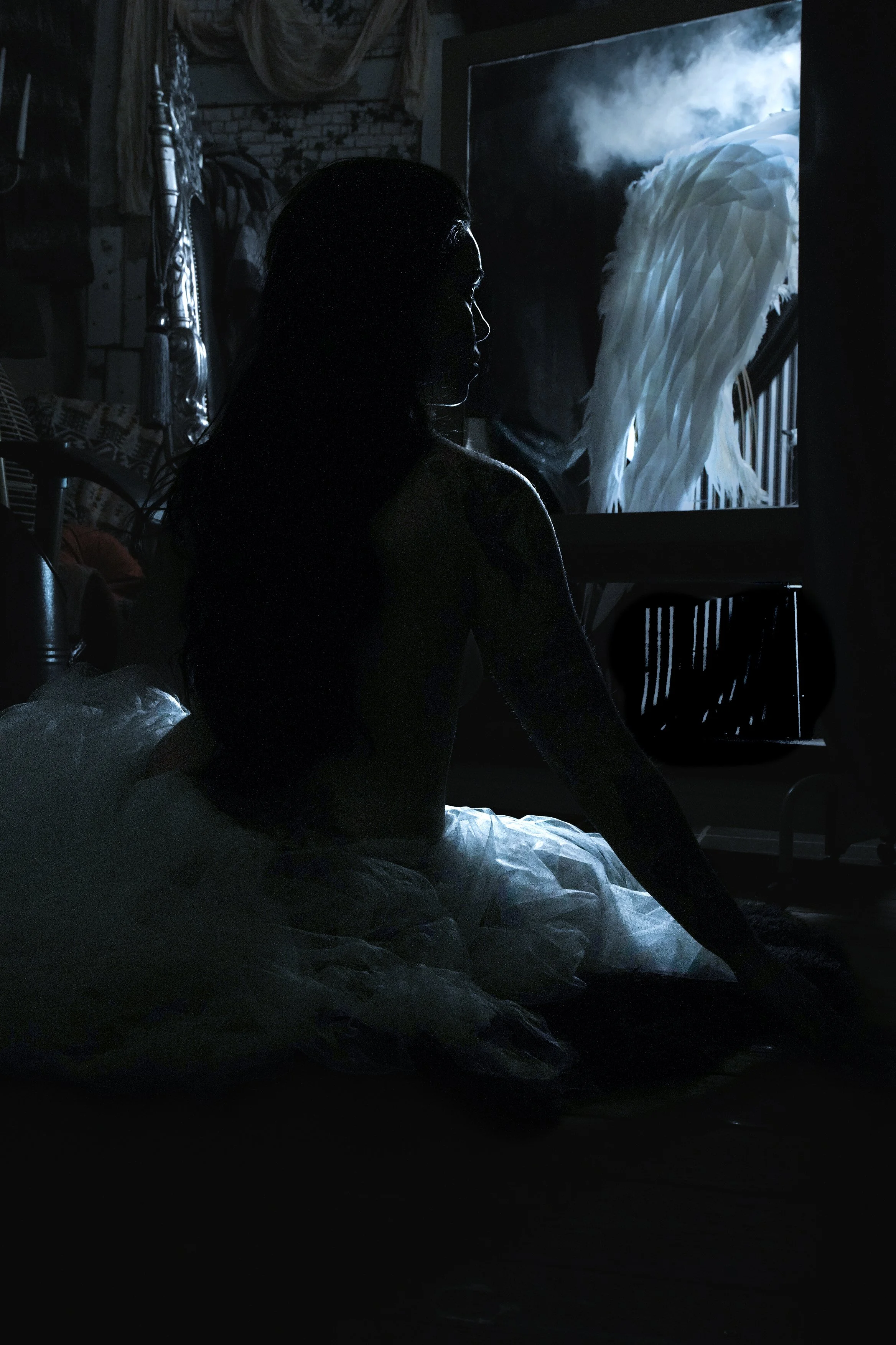 A woman sits on the floor in a dimly lit room with her side to the camera, looking at a window with angel wings hanging outside. The room has various items, including a chair and fabric drapes.