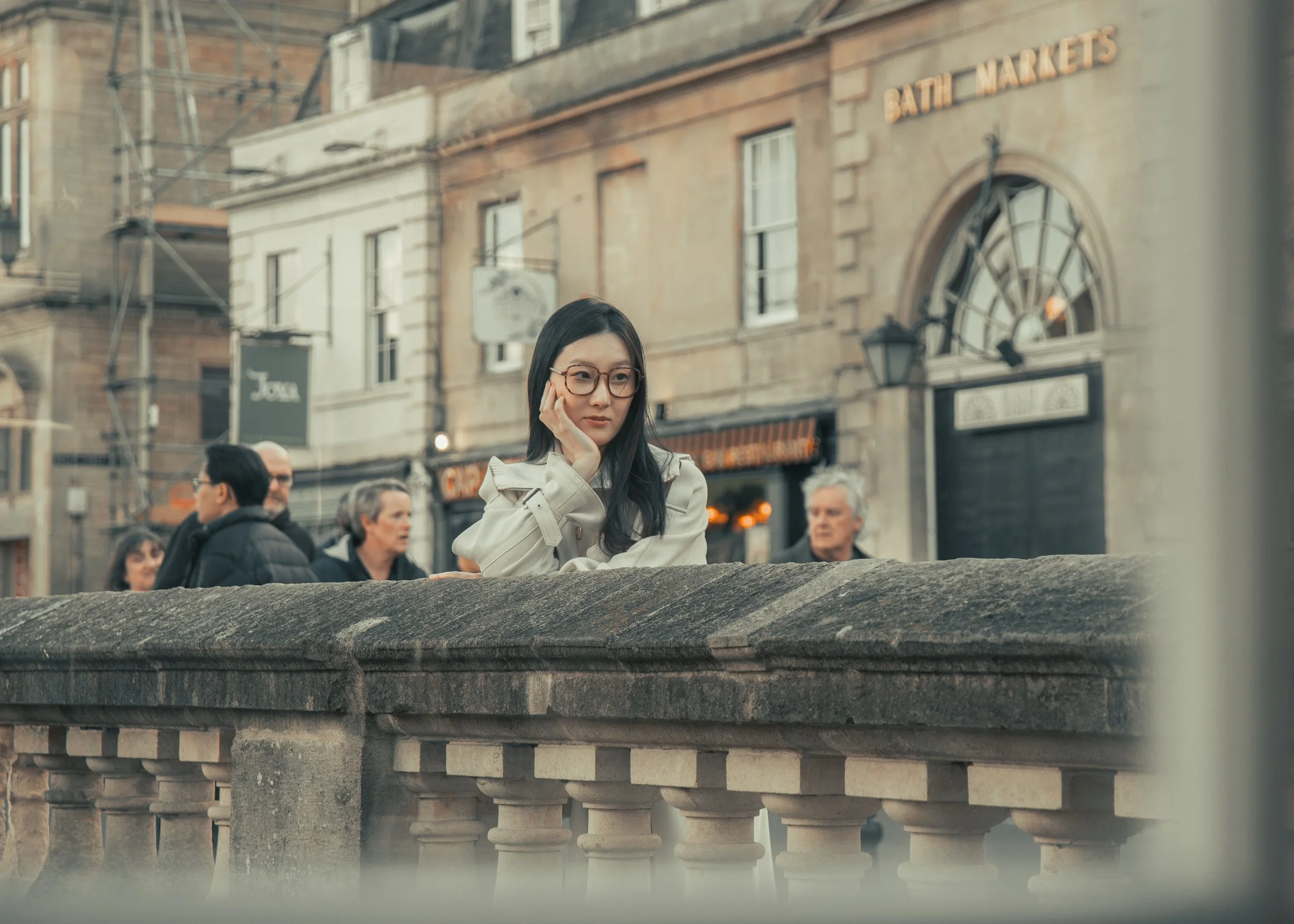 A young woman with glasses and a beige coat leaning on a stone railing in a city street, with old buildings and a sign reading 'Bath Markets' in the background.