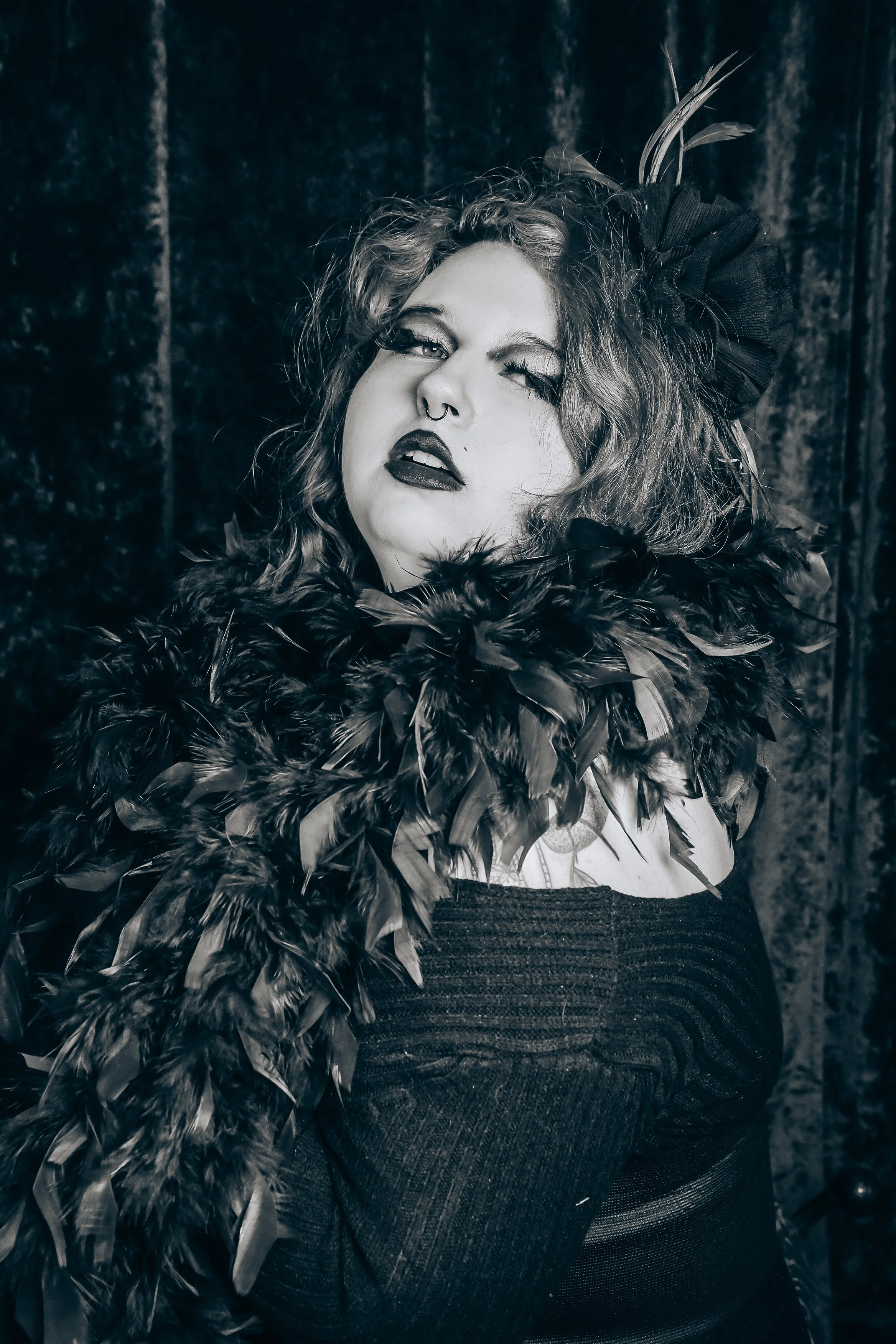 A woman with curly hair wearing dark lipstick, a septum piercing, and a feathered boa, posing against a dark textured background.
