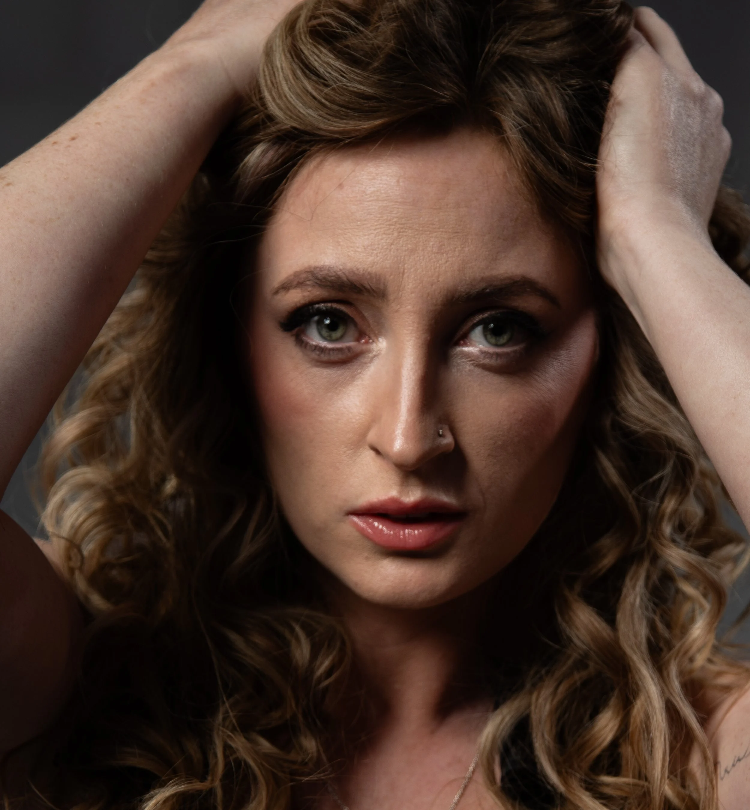 Close-up of a woman with curly brown hair, green eyes, and a nose piercing, holding her hair with both hands, looking directly at the camera.