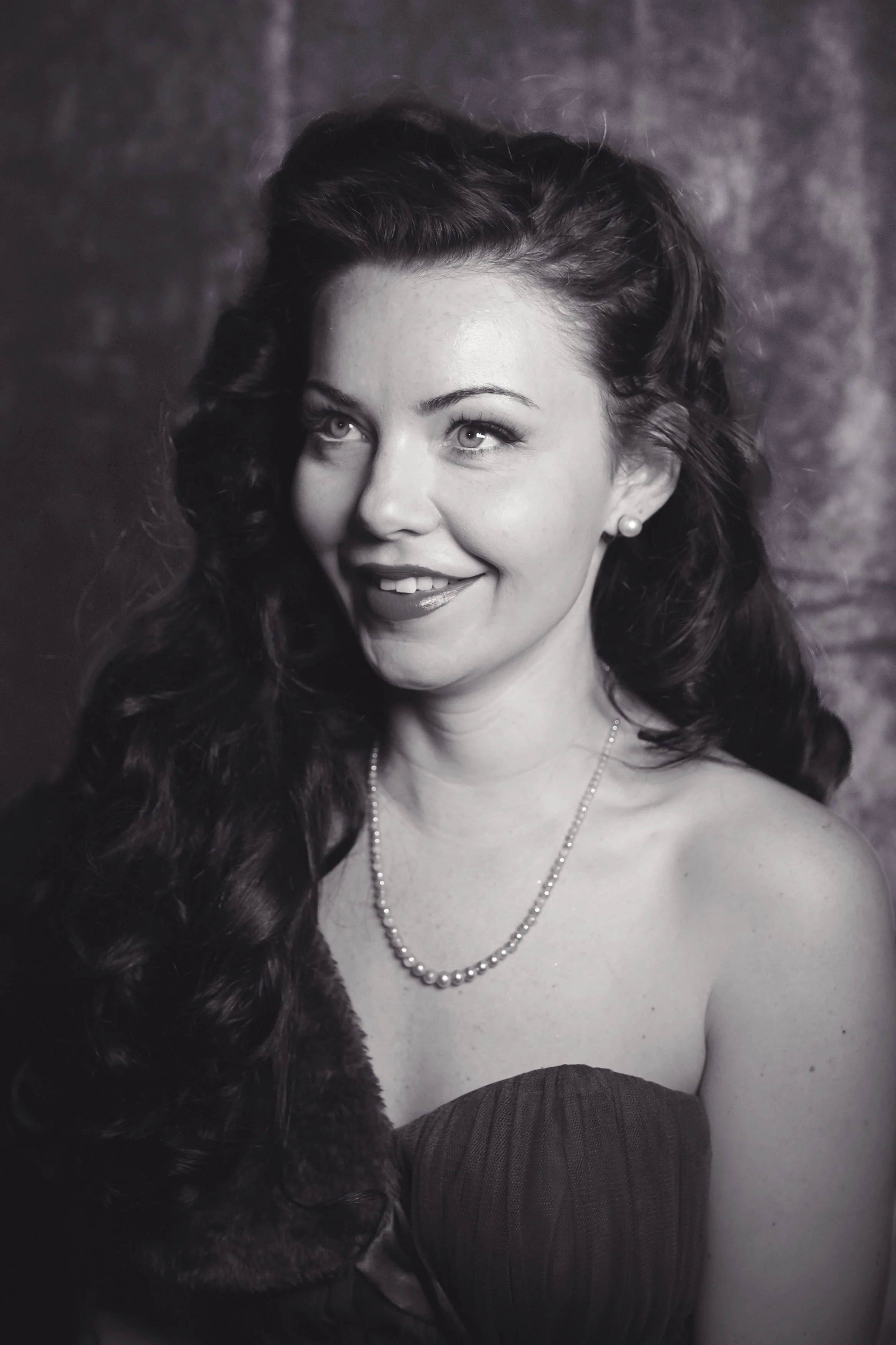 Black and white portrait of a woman with long, wavy dark hair, wearing a pearl necklace and earrings, and a strapless dress, smiling slightly and looking off to the side.