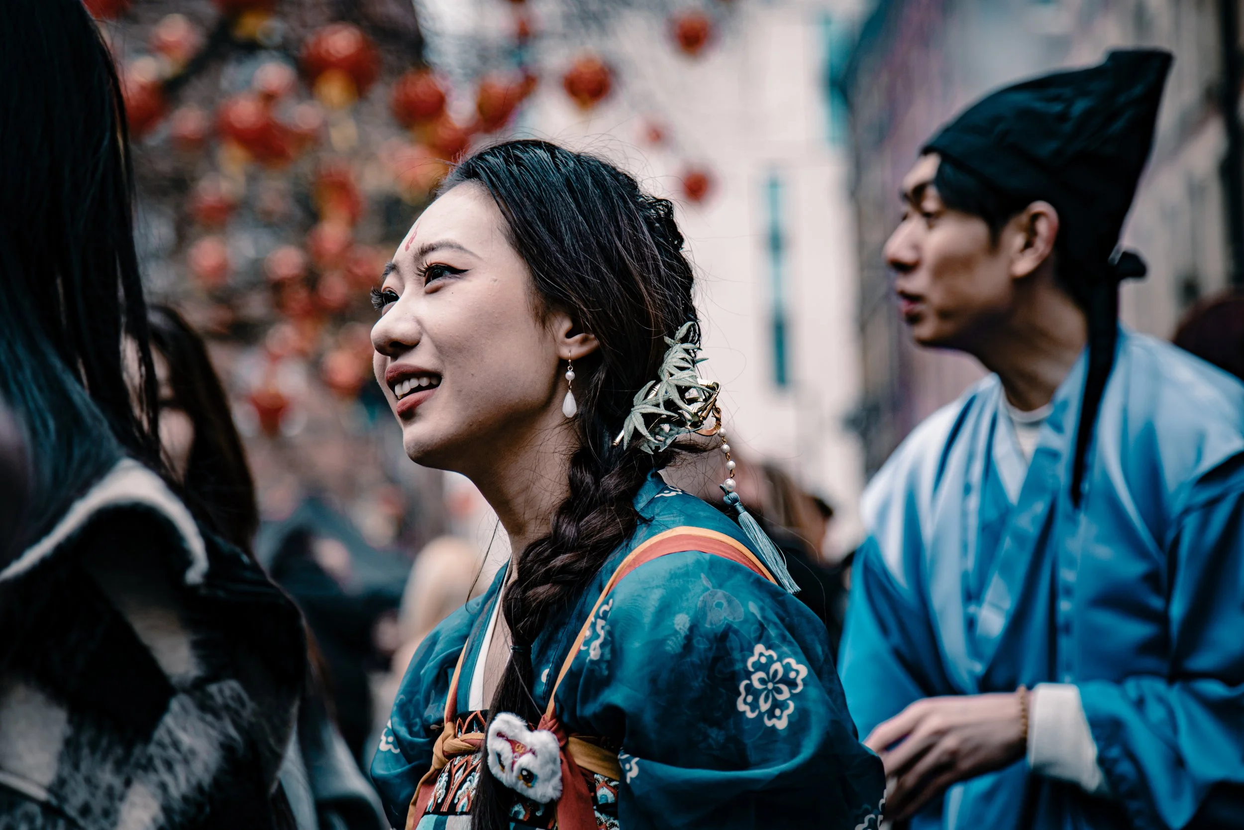 Two people in traditional Chinese clothing, one woman smiling and talking, and a man in a blue robe wearing a black hat, in a busy outdoor setting with blurred background of red lanterns and buildings.
