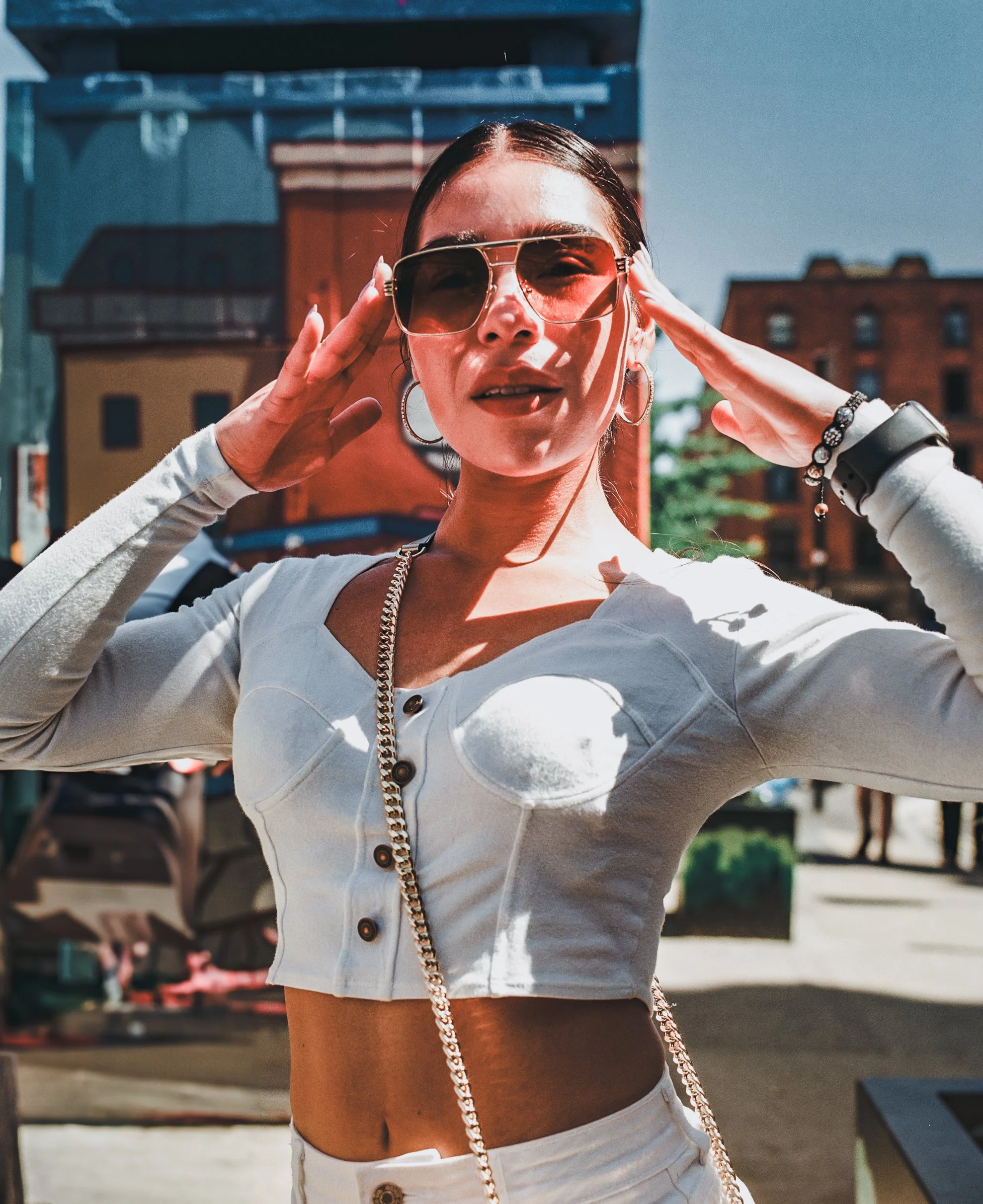 A woman wearing sunglasses, a white cropped jacket, and white pants, standing outdoors in sunlight, with buildings in the background.