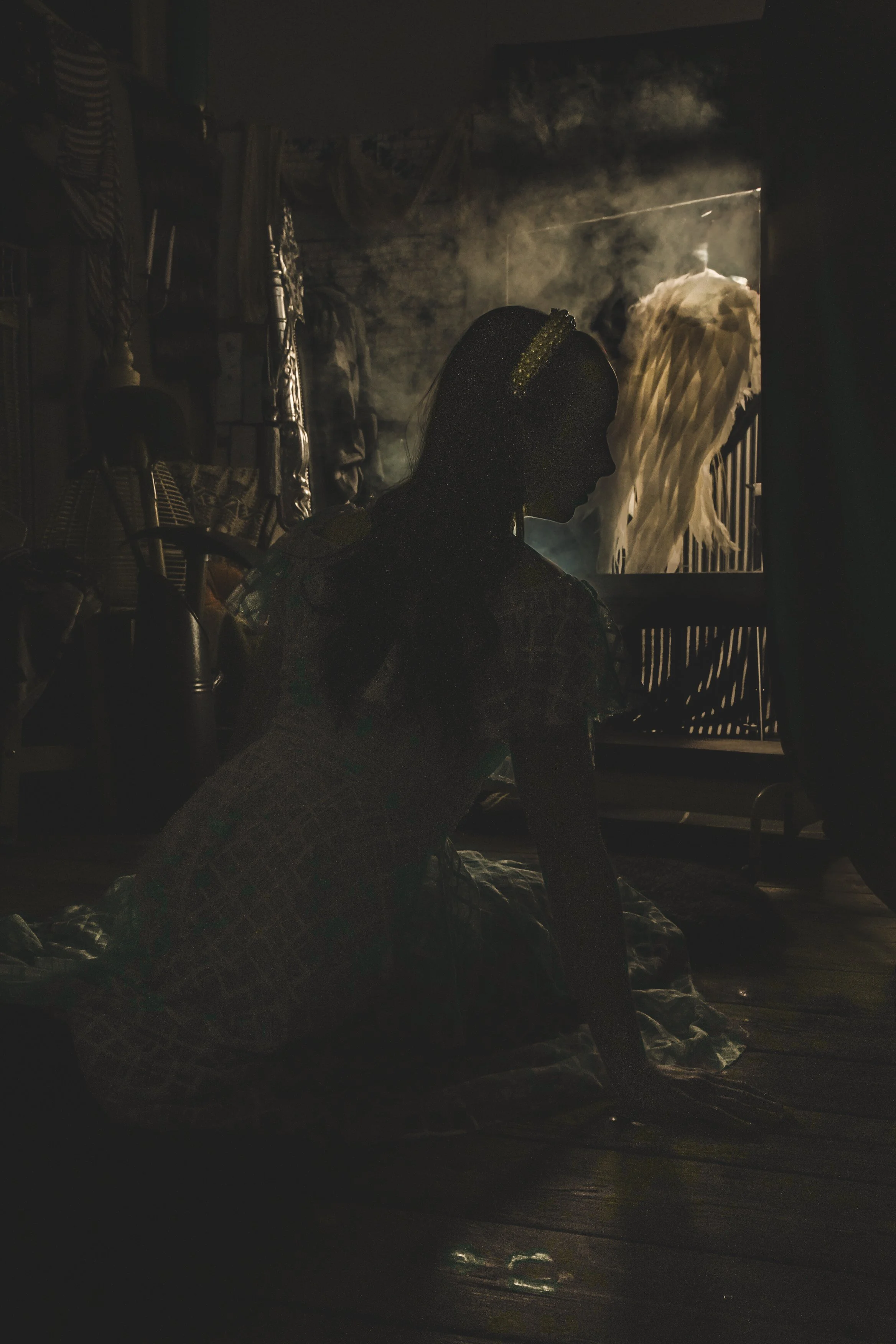 Silhouette of a woman sitting on the floor in a dimly lit room, with a large window revealing a figure in a white dress or costume in the bright light outside.