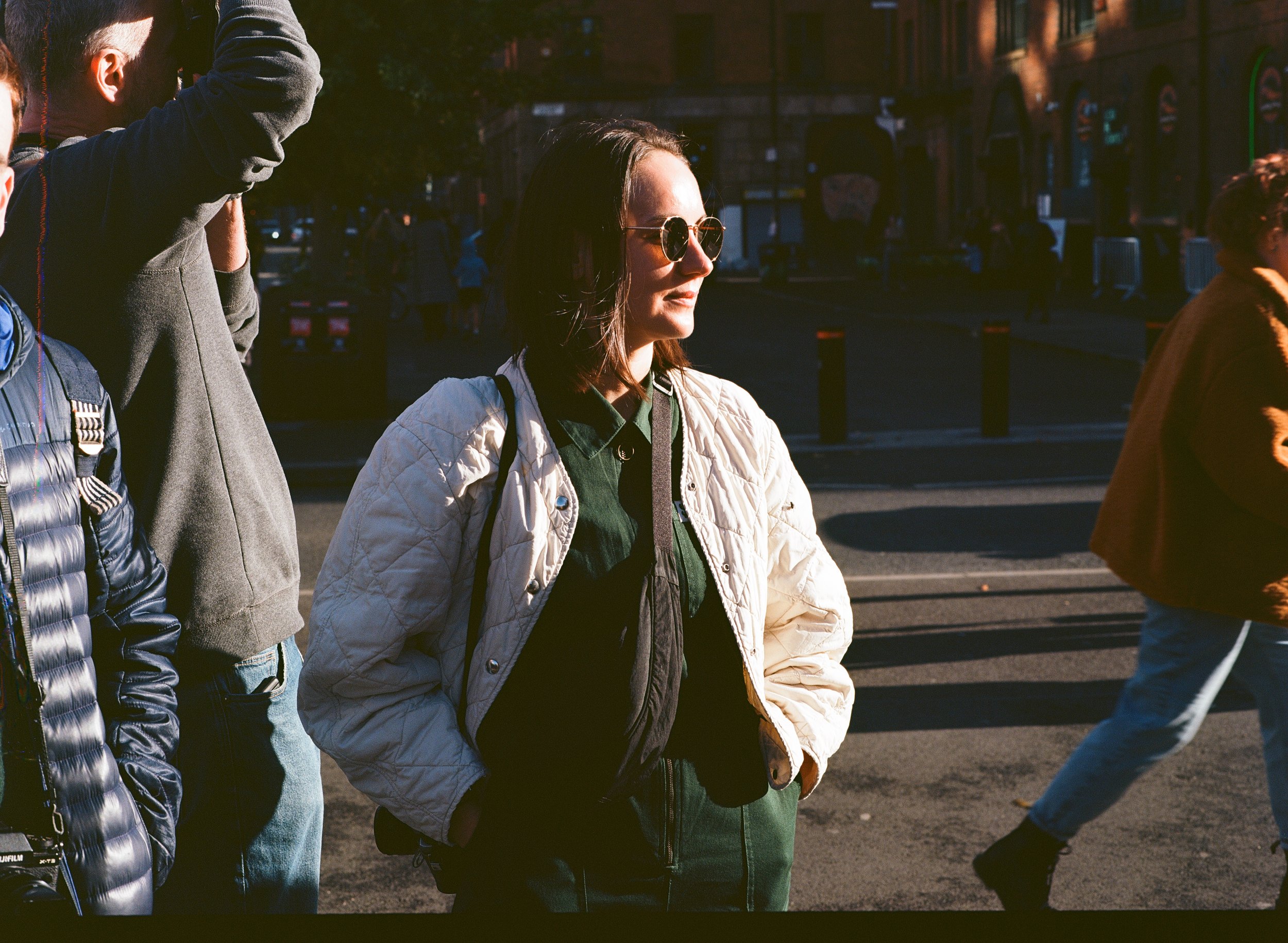 A woman with shoulder-length dark hair wearing sunglasses, a white quilted jacket, and a backpack standing in a city street during sunset.