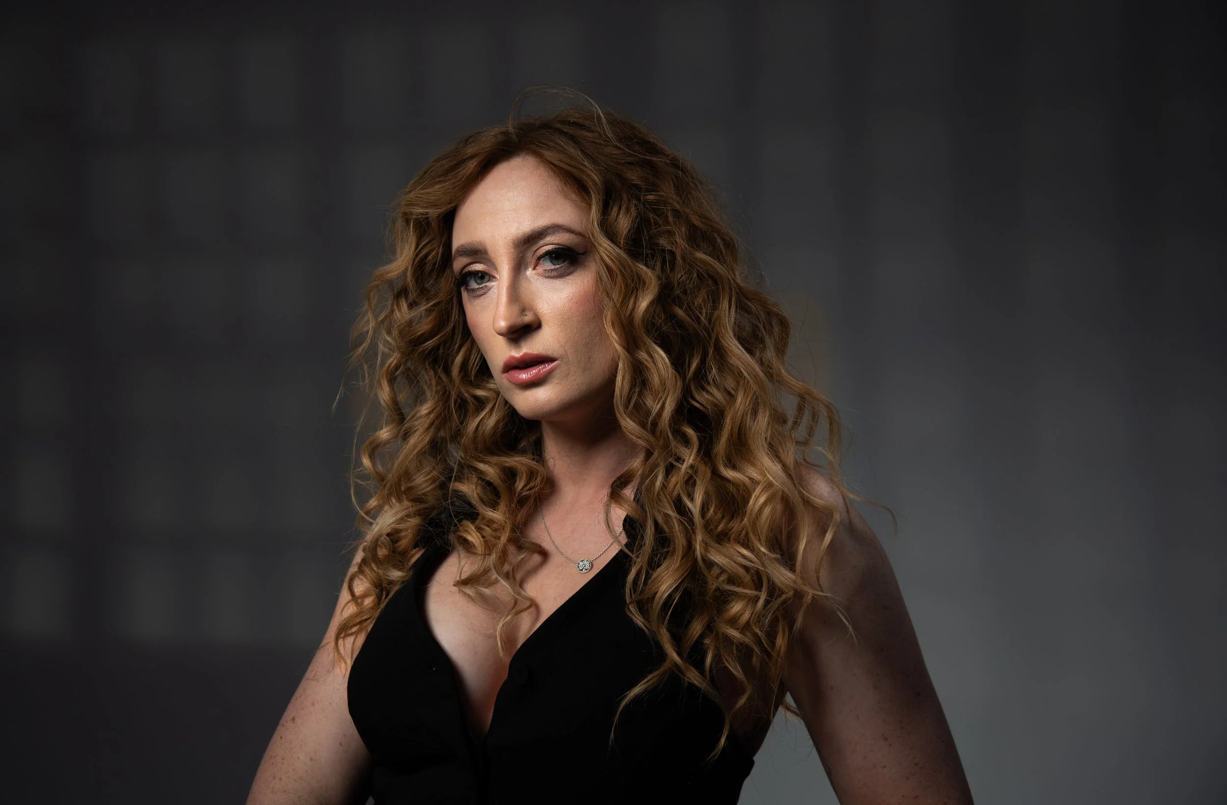 A woman with long, curly red hair wearing a black sleeveless top and a silver necklace poses against a dark background.