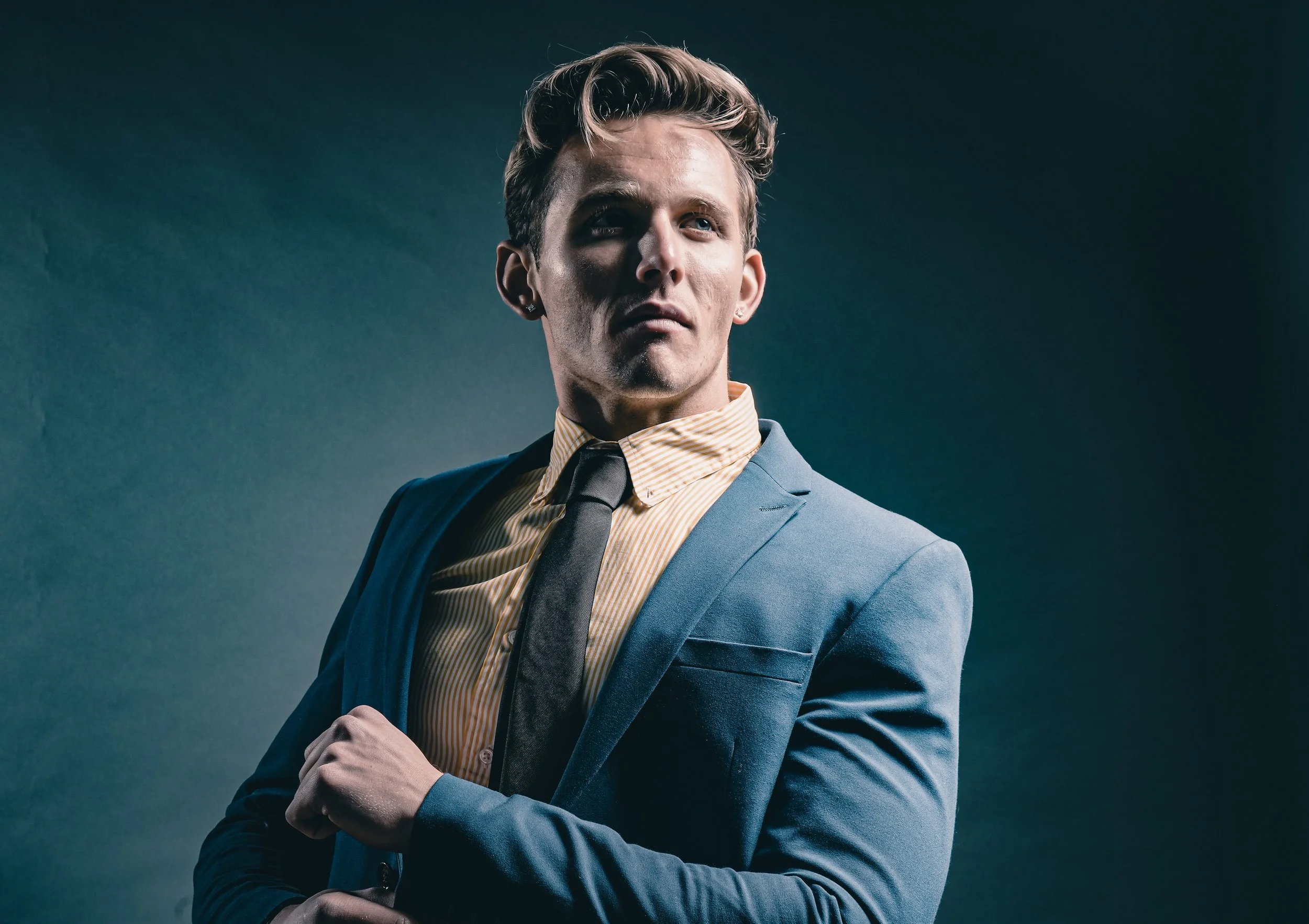 A young man in a light blue suit, yellow striped shirt, and black tie looking confidently to the side, against a dark background.