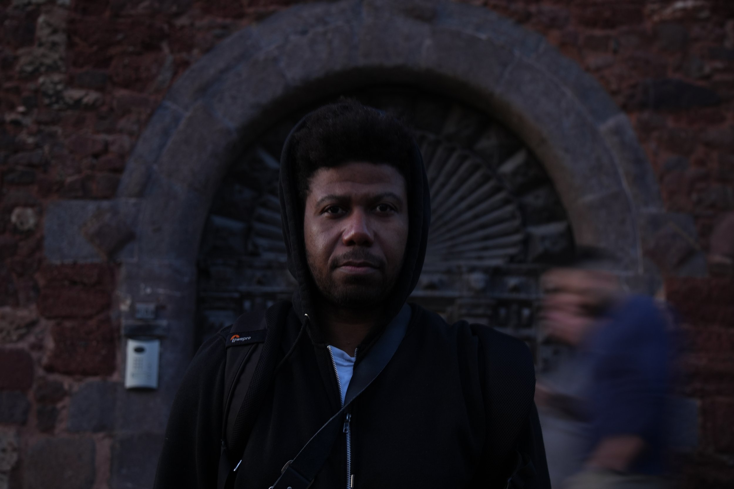 A man with dark hair and a beard standing in front of a stone archway with a decorative circular interior design. The man is wearing a black hoodie and a backpack, and there is a blurred figure walking past him on the right side.