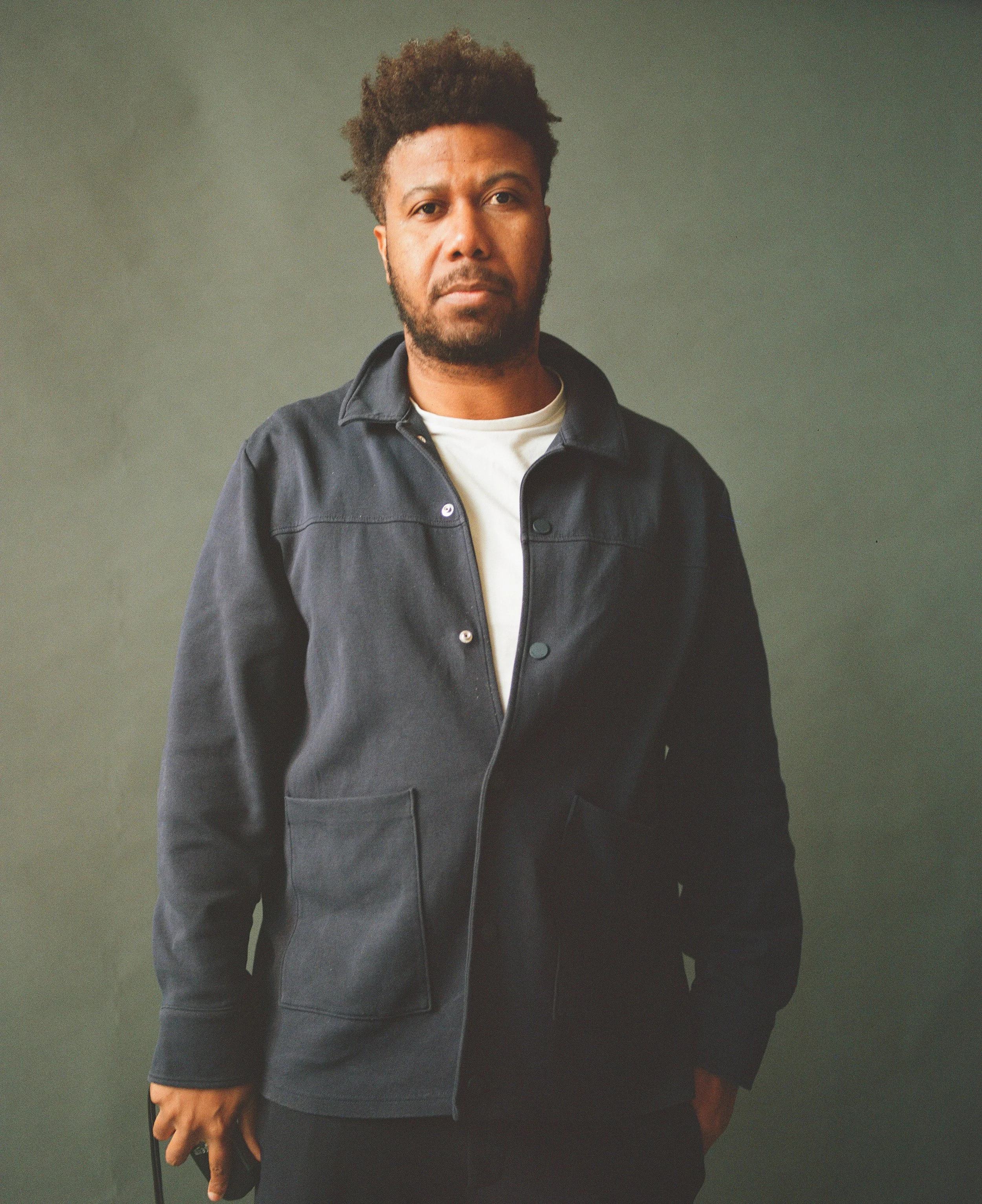 Portrait of a man with curly hair and beard wearing a black jacket and white shirt standing against a plain background.