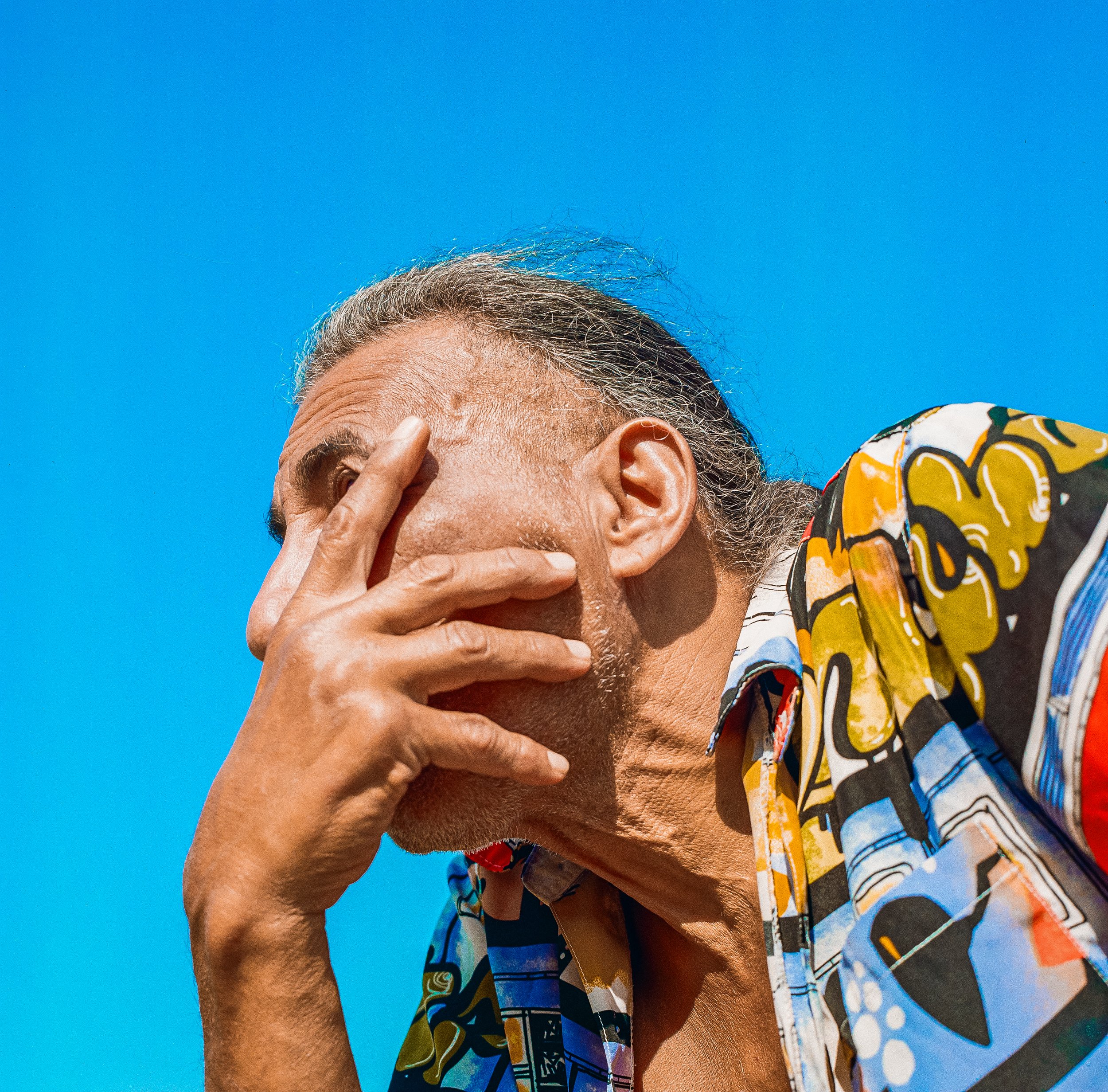 A man with gray hair and a colorful patterned shirt covering his face with his hand against a bright blue sky.