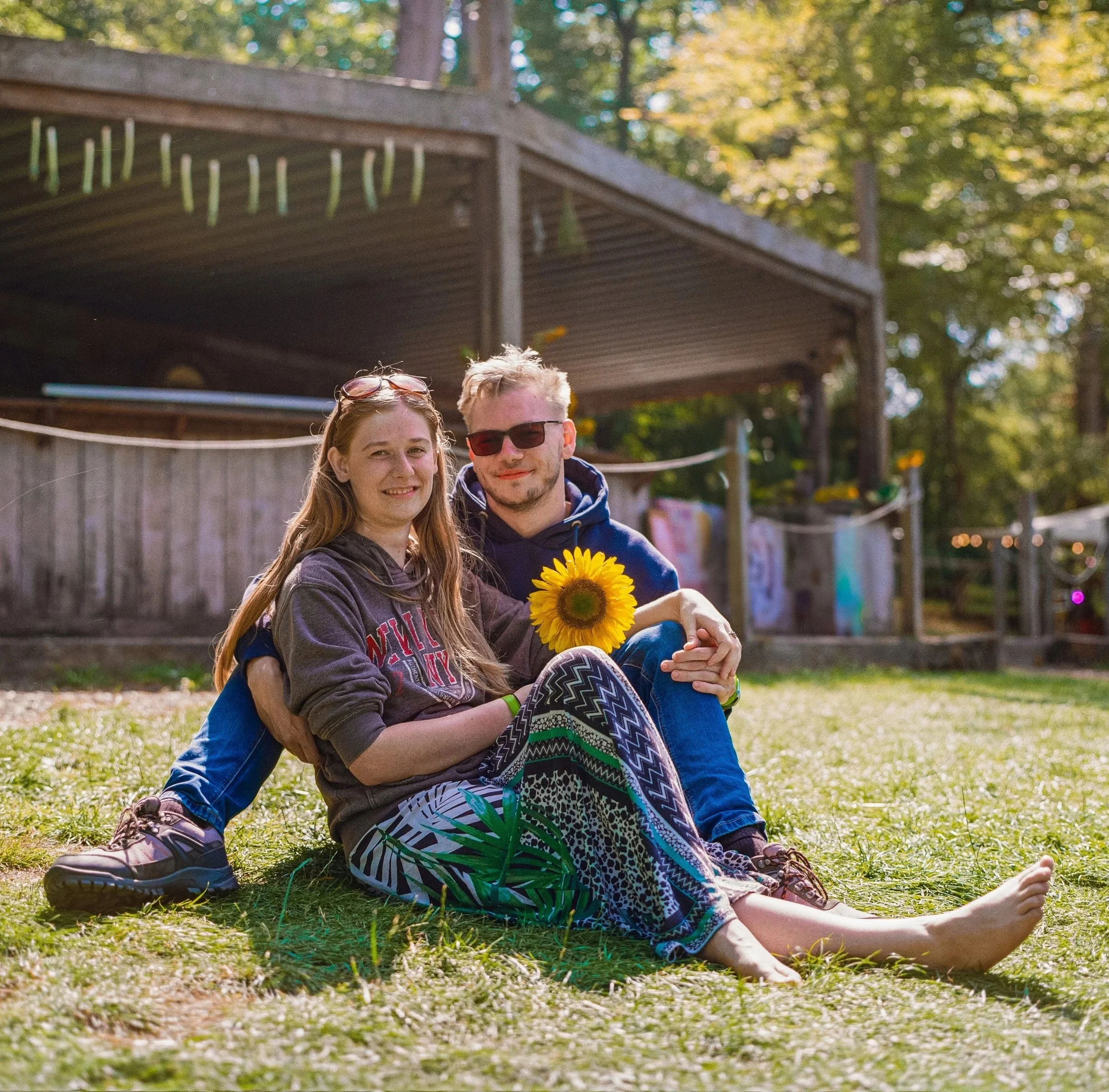 A young woman and a young man sitting on the grass outdoors, smiling at the camera, with the woman holding a sunflower. They are in a park or backyard setting with trees and a wooden structure in the background.