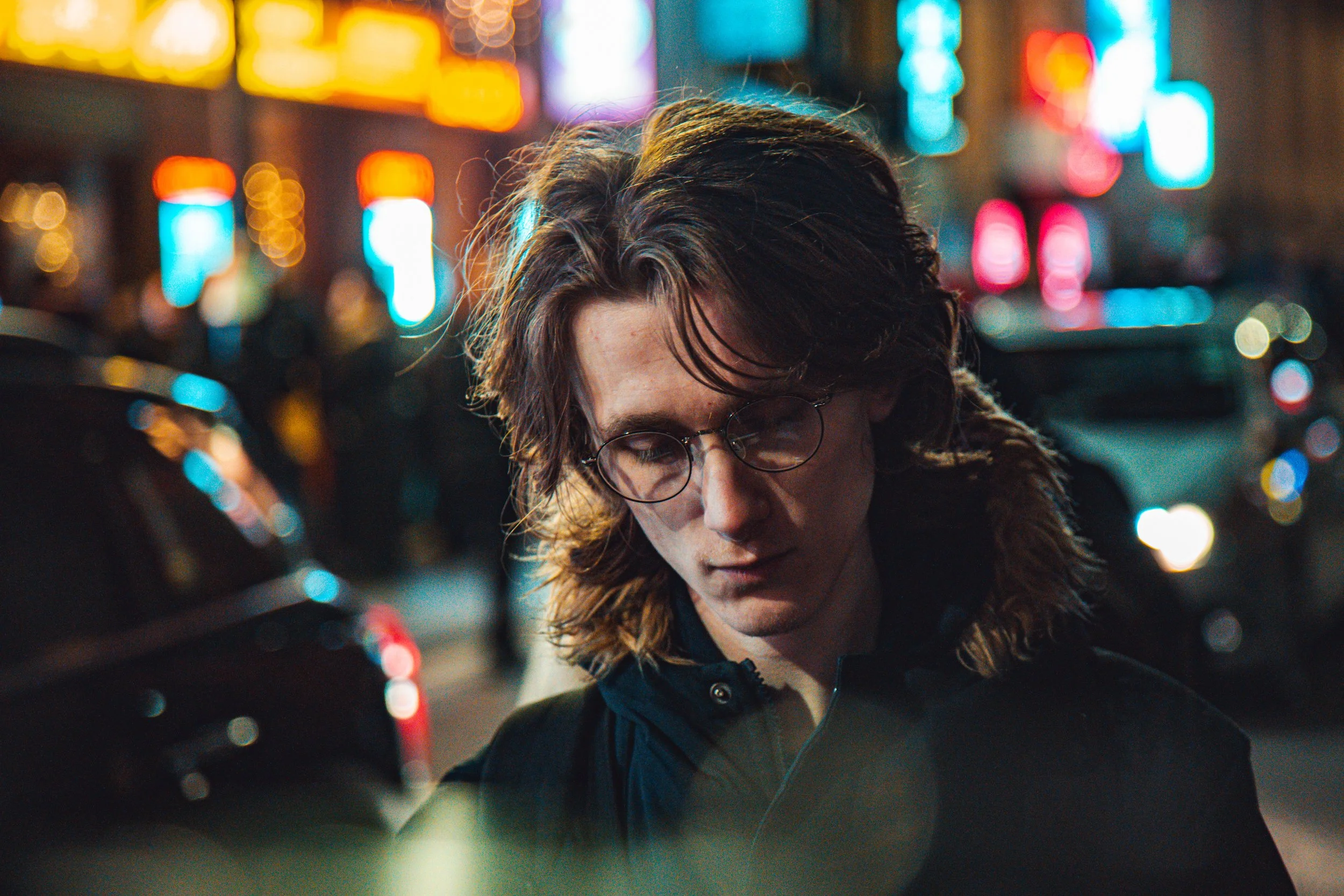 A young man with long, wavy brown hair and glasses standing on a busy city street at night, illuminated by colorful neon lights in the background.