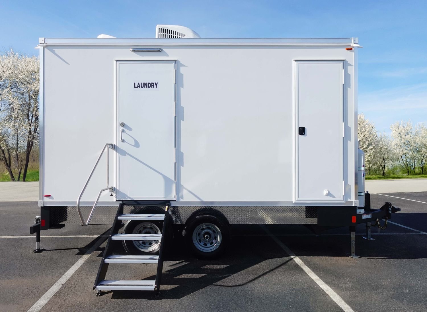 White mobile trailer with a door labeled 'Laundry', a small set of stairs, and a side door, parked in a lot with trees in the background.