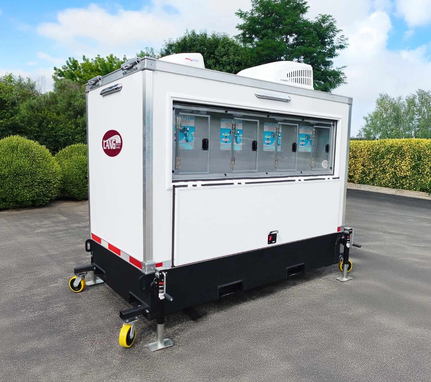 Mobile beverage vending trailer with a glass window display, branded with Lang logo, set on a paved lot with green shrubbery and trees in the background.