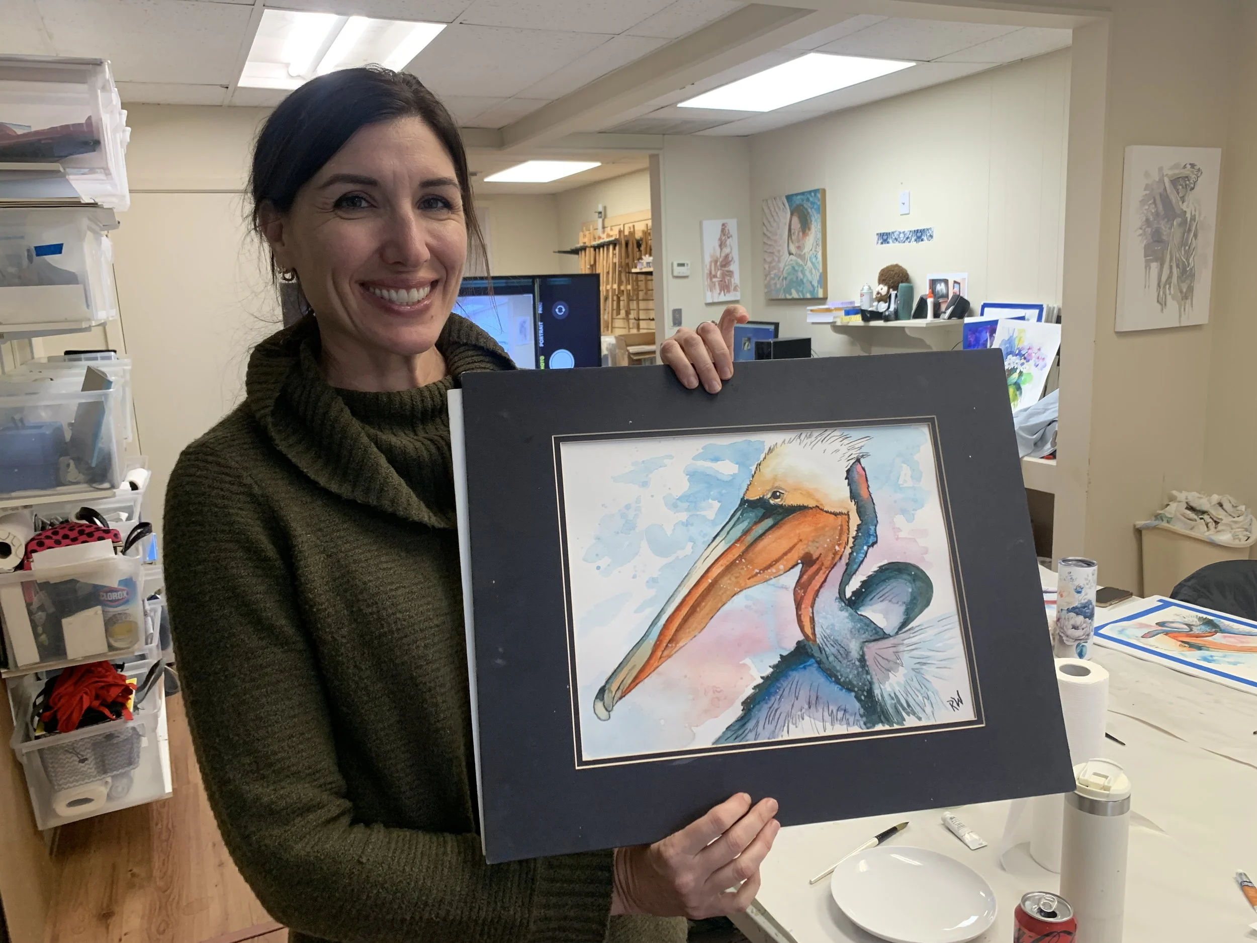 A woman with dark hair smiling and holding a framed watercolor painting of a pelican, in an art studio with paintings and art supplies in the background.