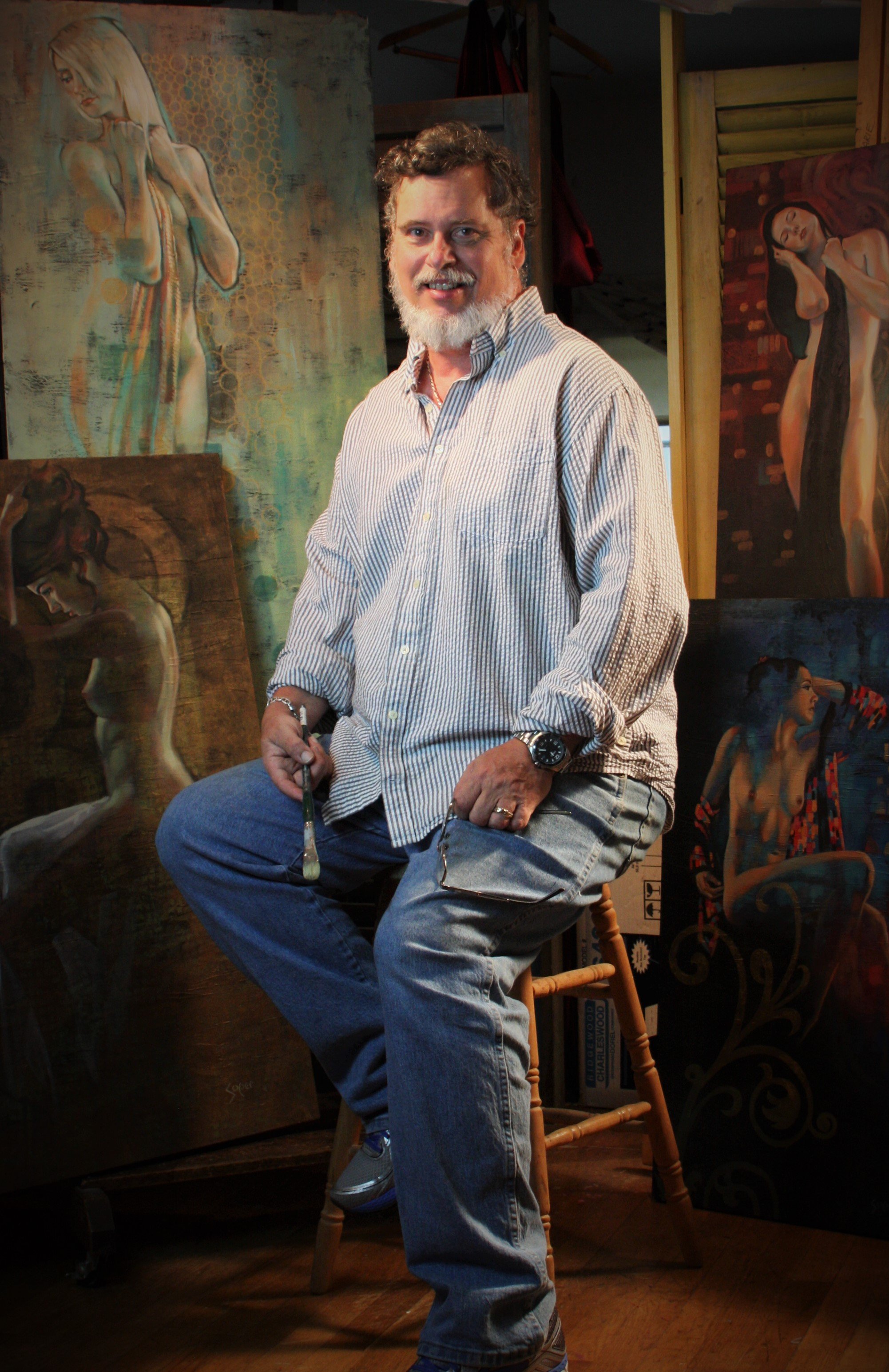 An artist sitting on a wooden stool surrounded by various paintings of women in an art studio.