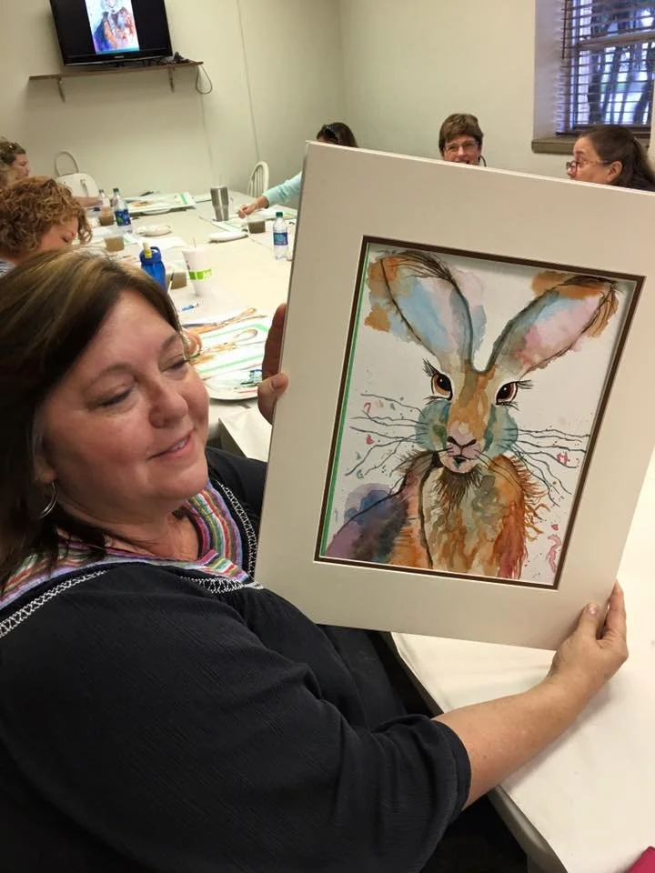 Woman holding a colorful watercolor painting of a rabbit with tall ears and detailed features, sitting at a table in a room with other people and a television on the wall.