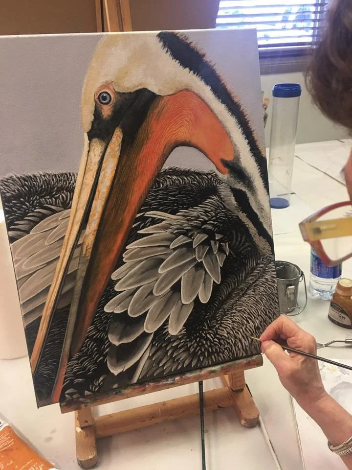 A person is painting a realistic detailed portrait of a pelican on a canvas. The artist is using a fine brush, and the painting depicts the pelican's beak, eye, and feathers with intricate shading and color variations.