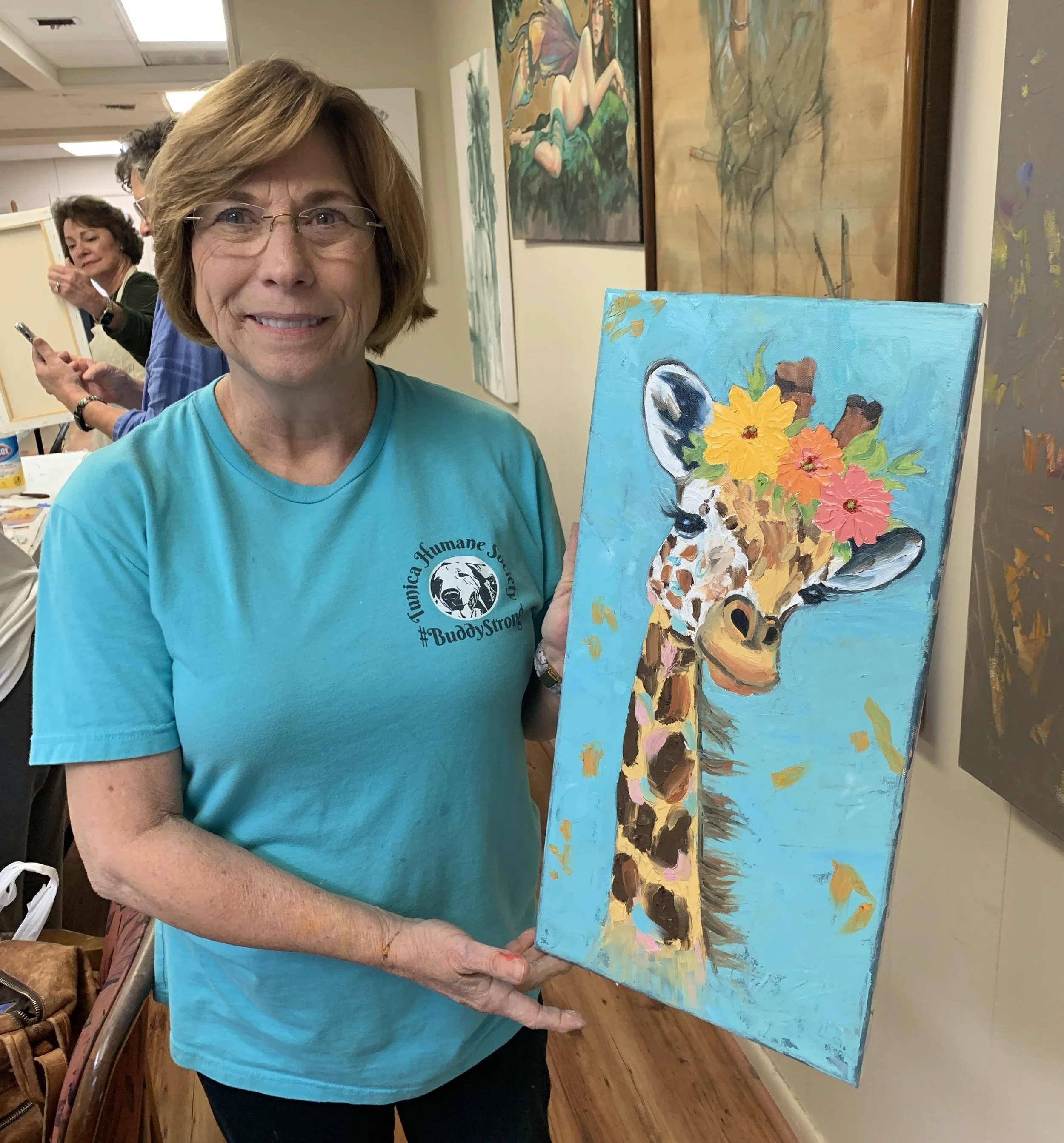 A woman holding a colorful painting of a giraffe with flowers on its head, in an art gallery or studio.